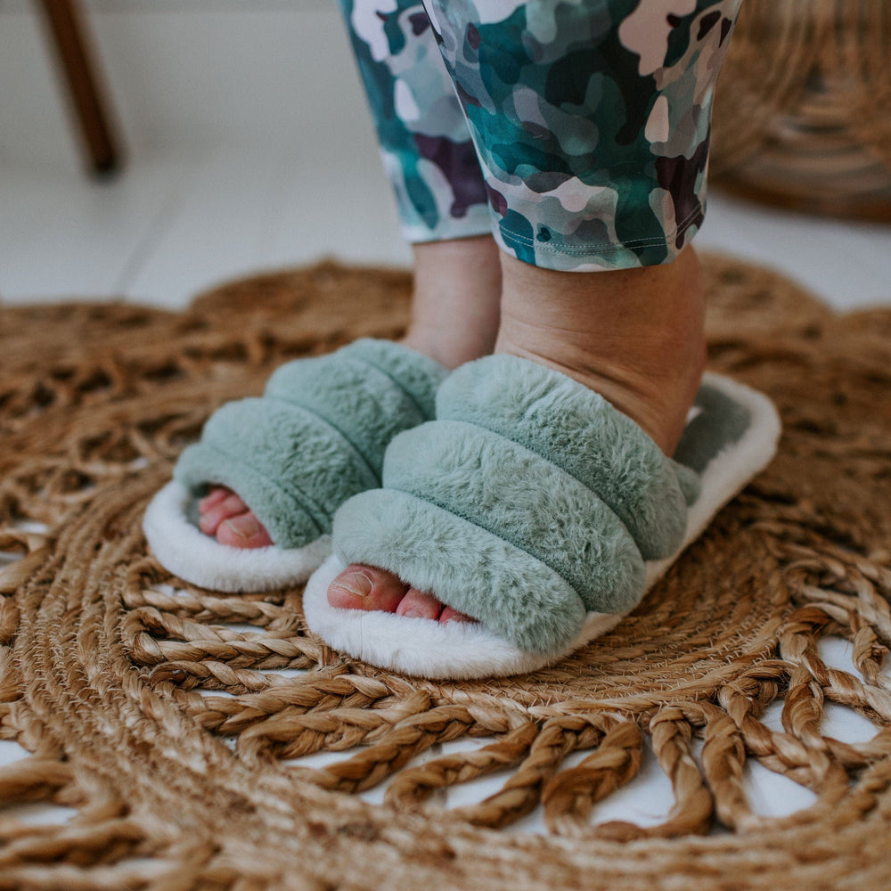 A person wearing green fuzzy slippers with rubber soles and camouflage pants stands on a woven rug.