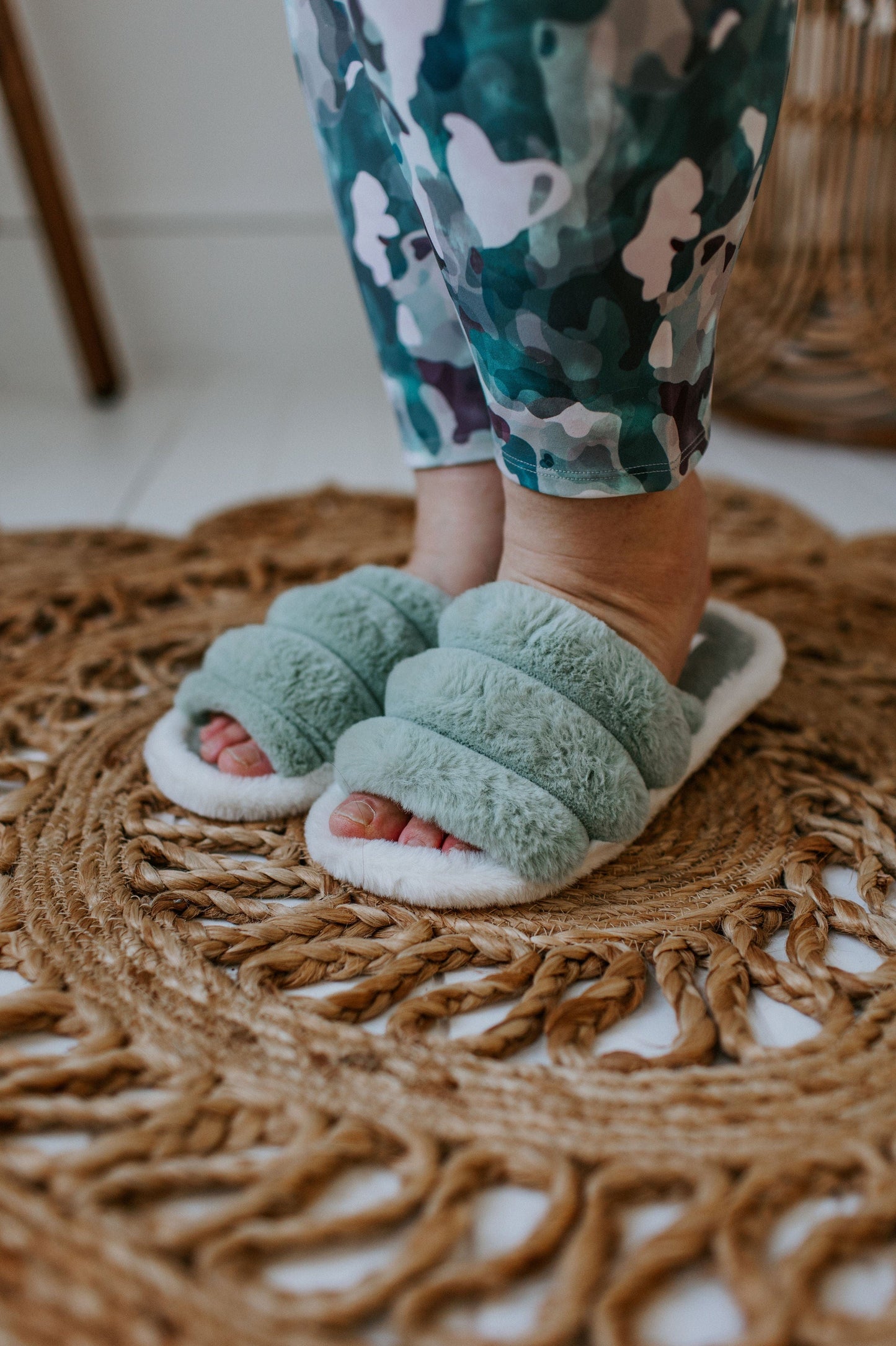 A person wearing green fuzzy slippers with rubber soles and camouflage pants stands on a woven rug.