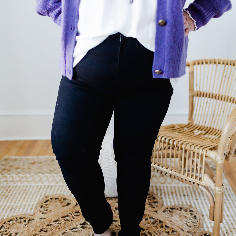 A person in a purple cardigan and high-rise black pants stands barefoot on a woven rug next to a rattan chair.