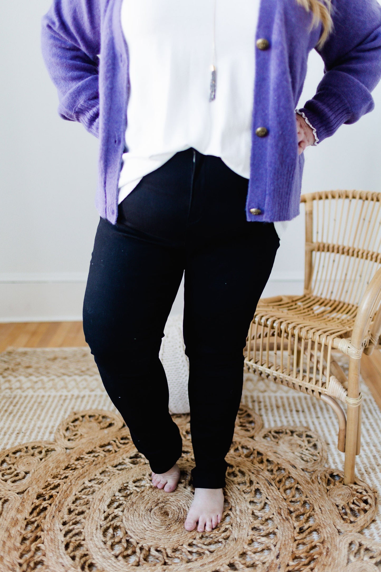 A person in a purple cardigan and high-rise black pants stands barefoot on a woven rug next to a rattan chair.