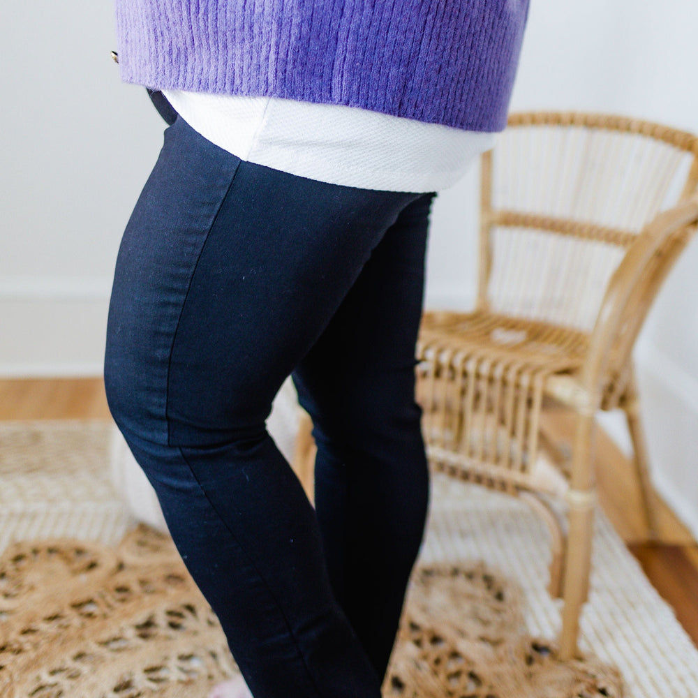 A person wearing a purple sweater and black skinny jeans stands on a woven rug beside a wicker chair.
