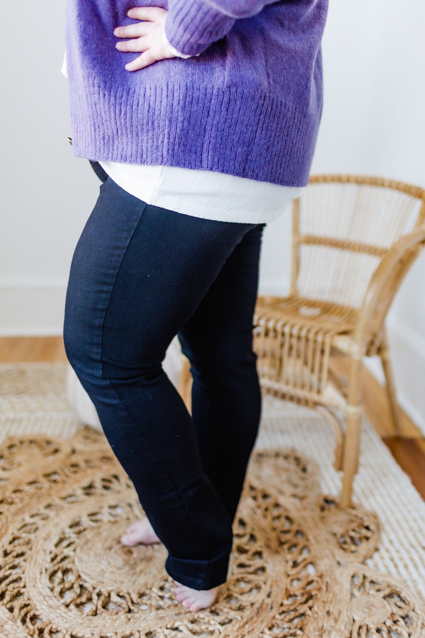 A person wearing a purple sweater and black skinny jeans stands on a woven rug beside a wicker chair.