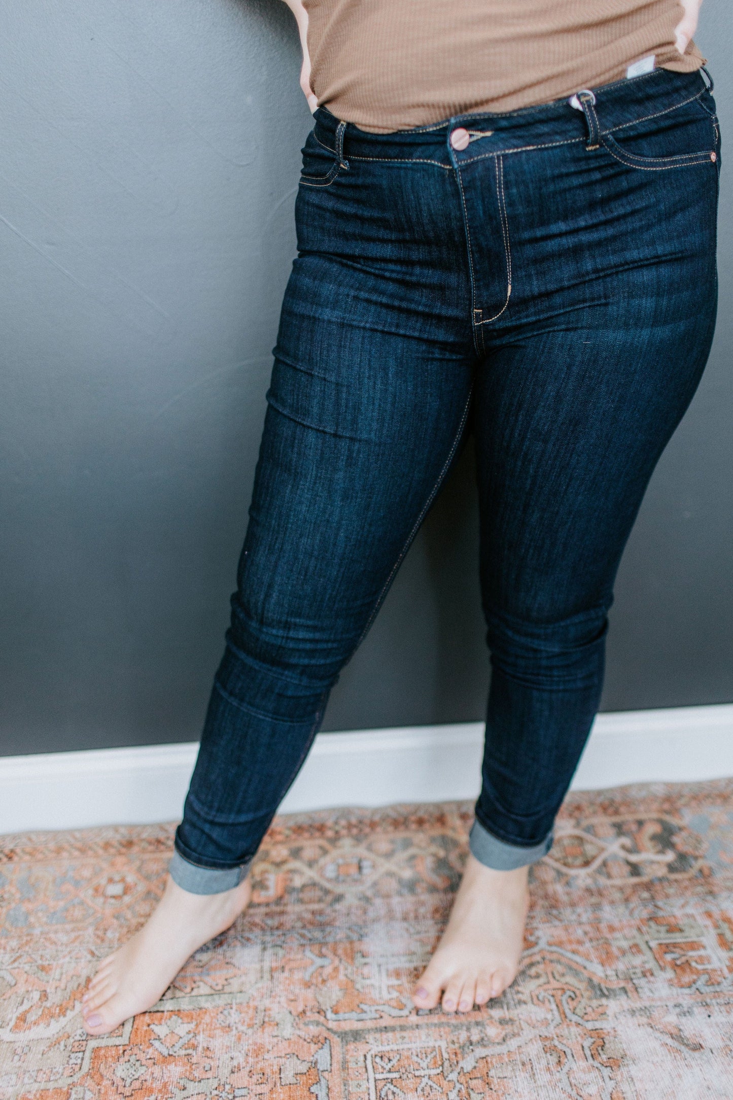 A person wearing rolled-up, high-rise dark denim jeans stands barefoot on a patterned rug against a gray wall.