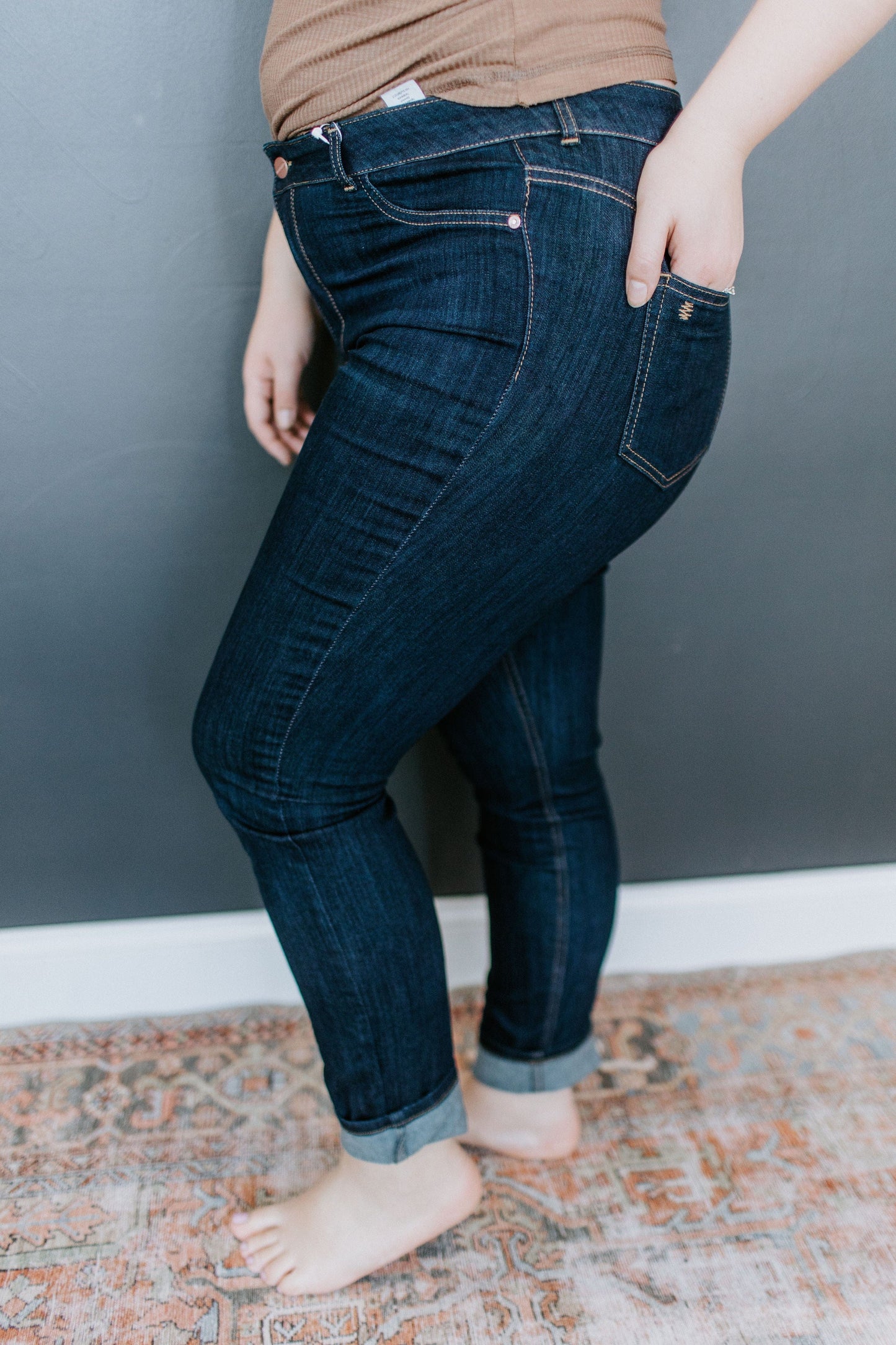 A person wearing high-rise dark denim jeans with rolled cuffs stands barefoot on a patterned carpet in front of a gray wall.