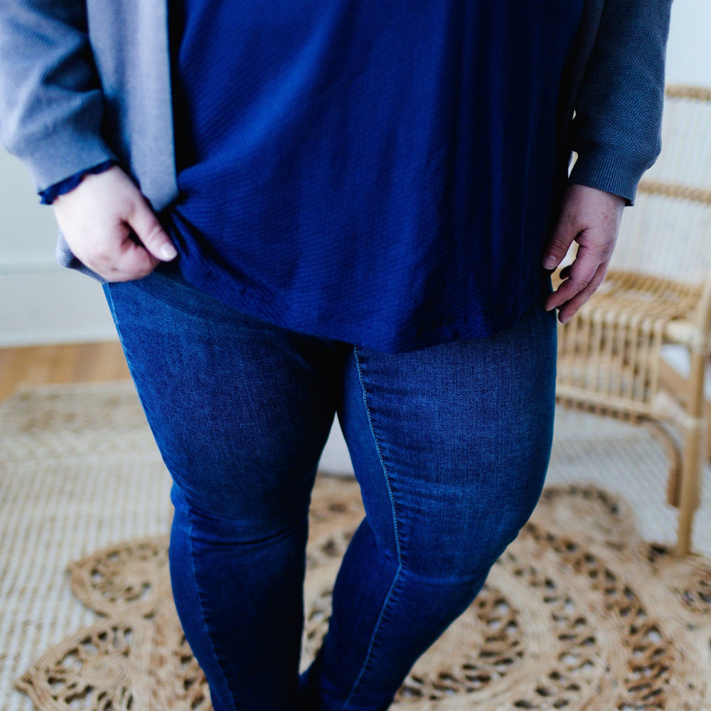 A person wearing the HIGH-RISE SKINNY JEGGING IN INDIGO from Love Marlow, paired with a navy top, is standing on a woven rug next to a wicker chair, shown from the shoulders down.