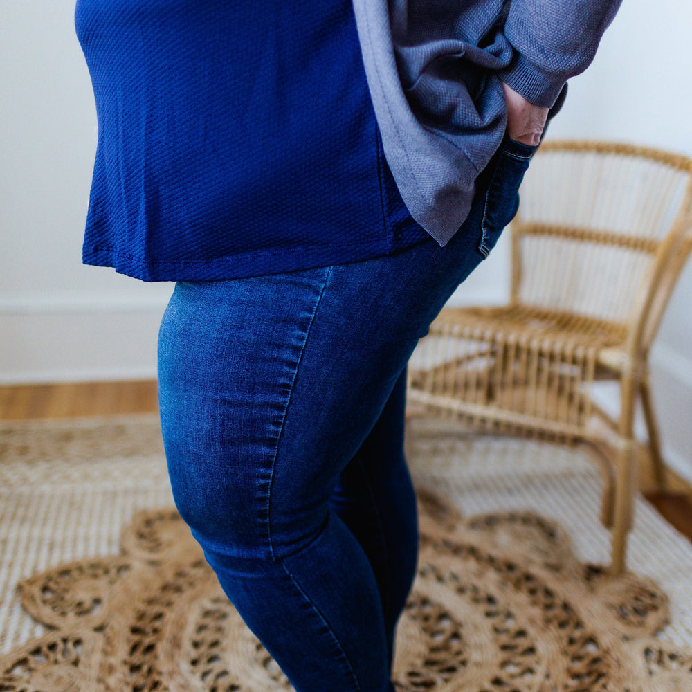 A person wearing a blue top and Love Marlow's HIGH-RISE SKINNY JEGGING IN INDIGO stands barefoot on a woven rug next to a wicker chair.