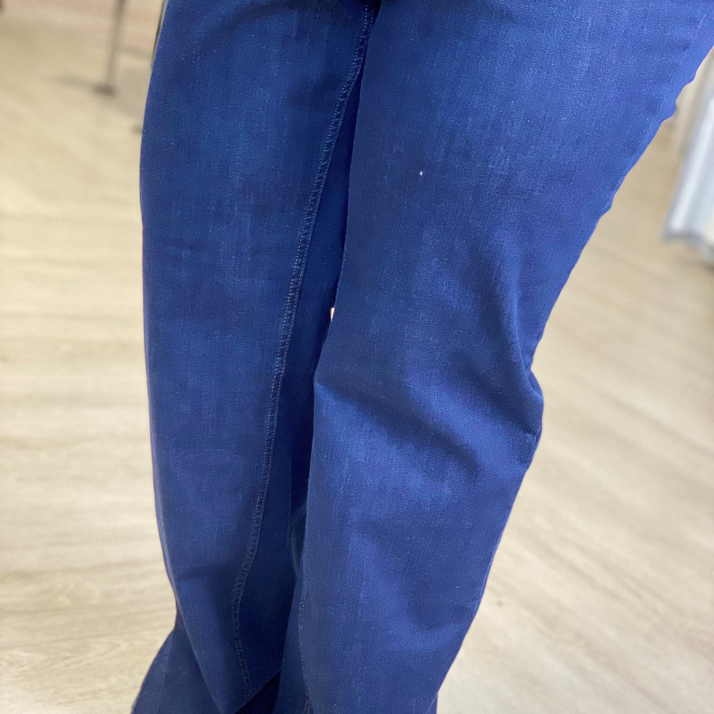 A person wearing high-rise, super stretchy blue jeans and a light green top stands indoors on a light wood floor.