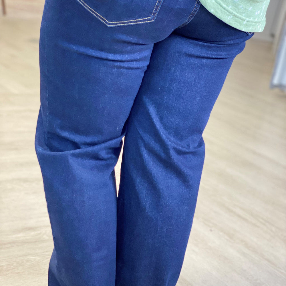 A person wearing high-rise, dark blue jeans and a green top is shown from the back, standing on a light-colored floor. The super stretchy material ensures comfort and a flattering fit.