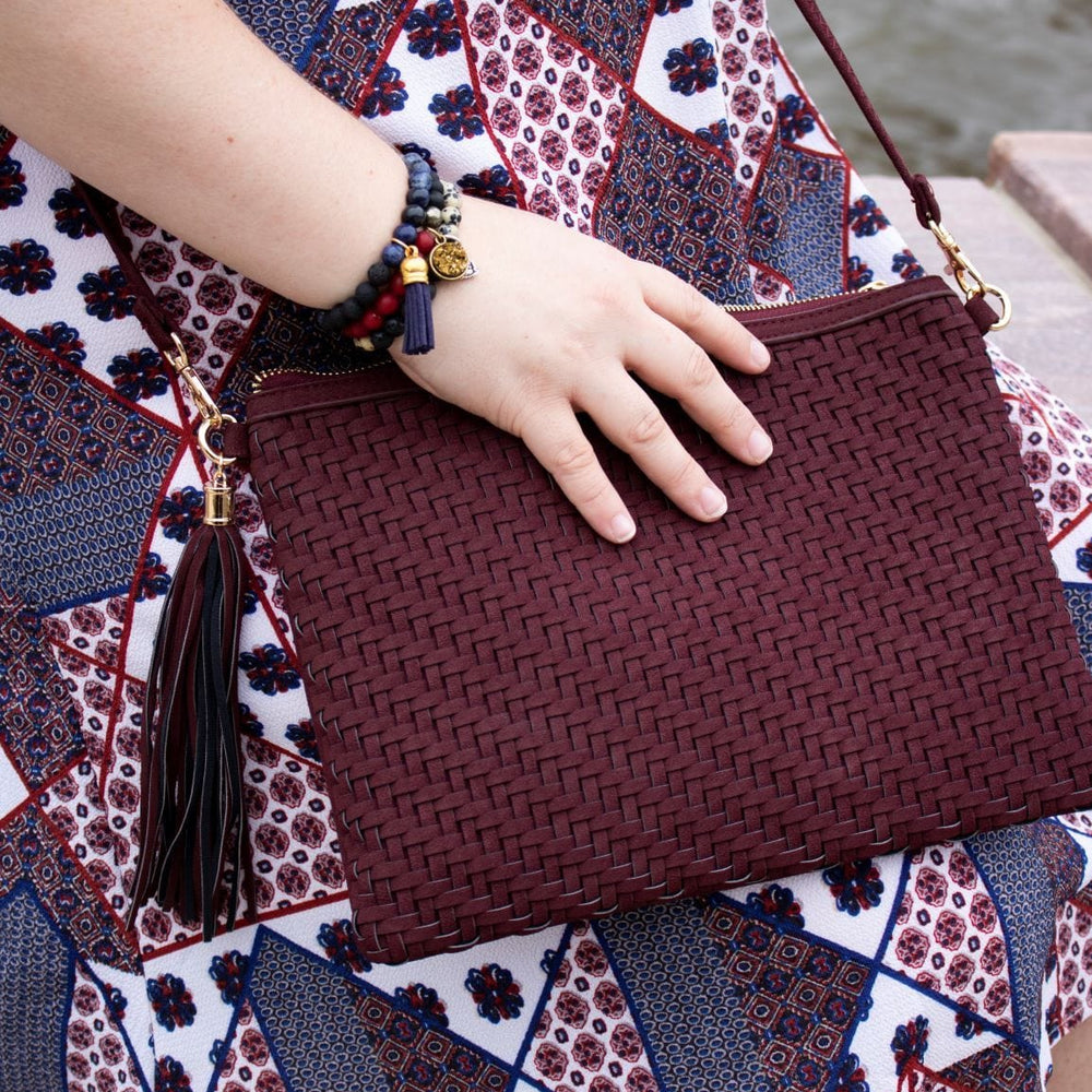 Sitting by the water, a person cradles a maroon woven clutch. They're adorned in a patterned dress, beaded bracelets, and beige shoes, capturing a serene moment with effortless style.