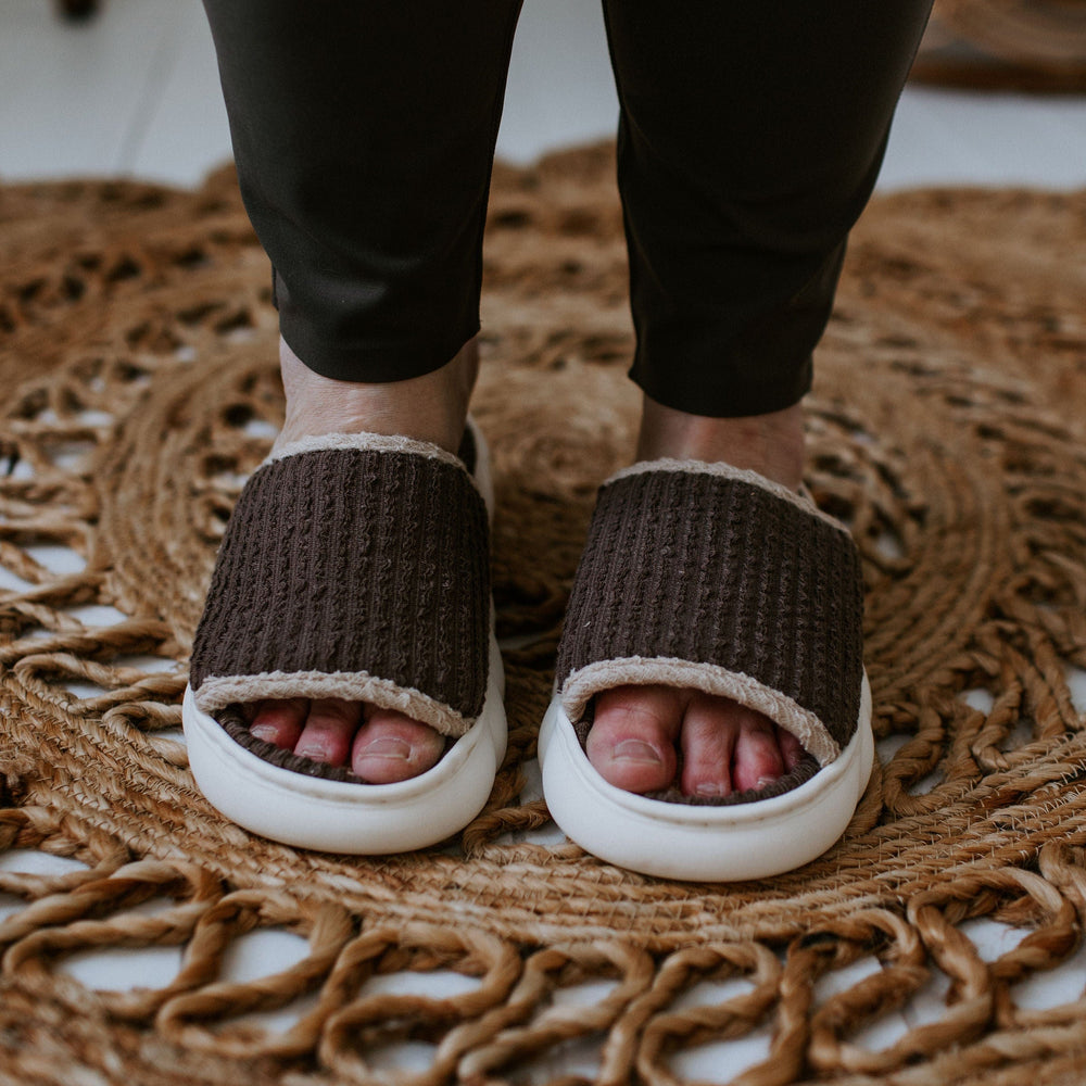 Close-up of a person wearing spa-inspired slides with dark brown knit fabric and white soles, standing on a woven circular rug.