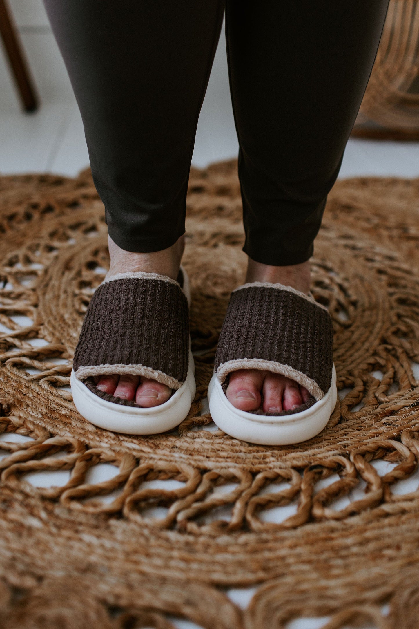 Close-up of a person wearing spa-inspired slides with dark brown knit fabric and white soles, standing on a woven circular rug.