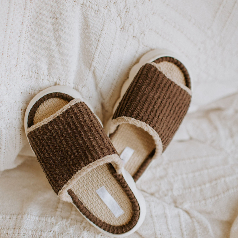 Brown ribbed slippers with white soles rest on a textured white fabric background, offering a spa-inspired slide experience.