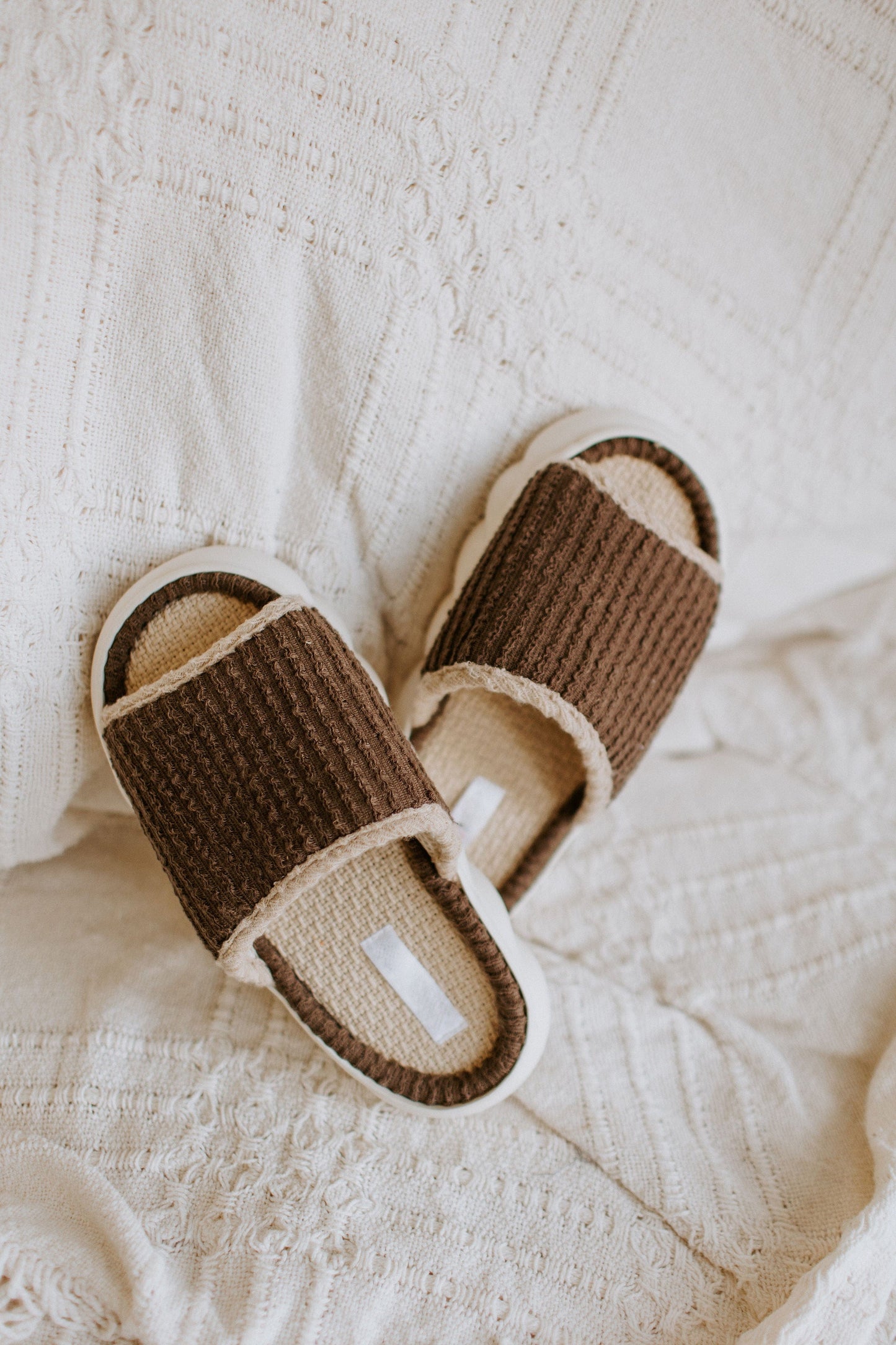 Brown ribbed slippers with white soles rest on a textured white fabric background, offering a spa-inspired slide experience.