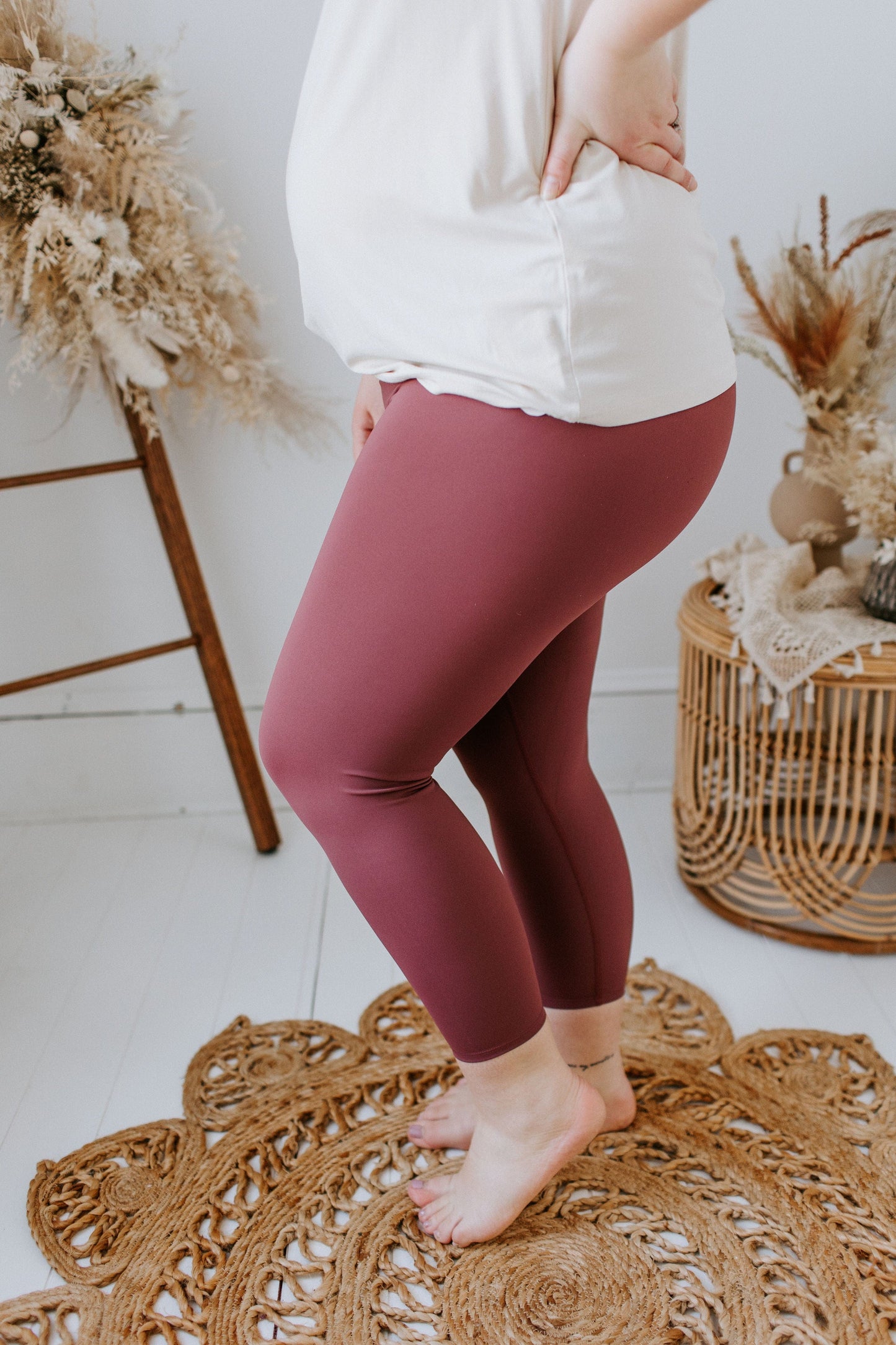 A person wearing a white top and Spanx© Booty Boost Active 7/8 Leggings in Midnight Rose, designed by Love Marlow with a contoured waistband, stands on a woven rug with dried plants in the background.