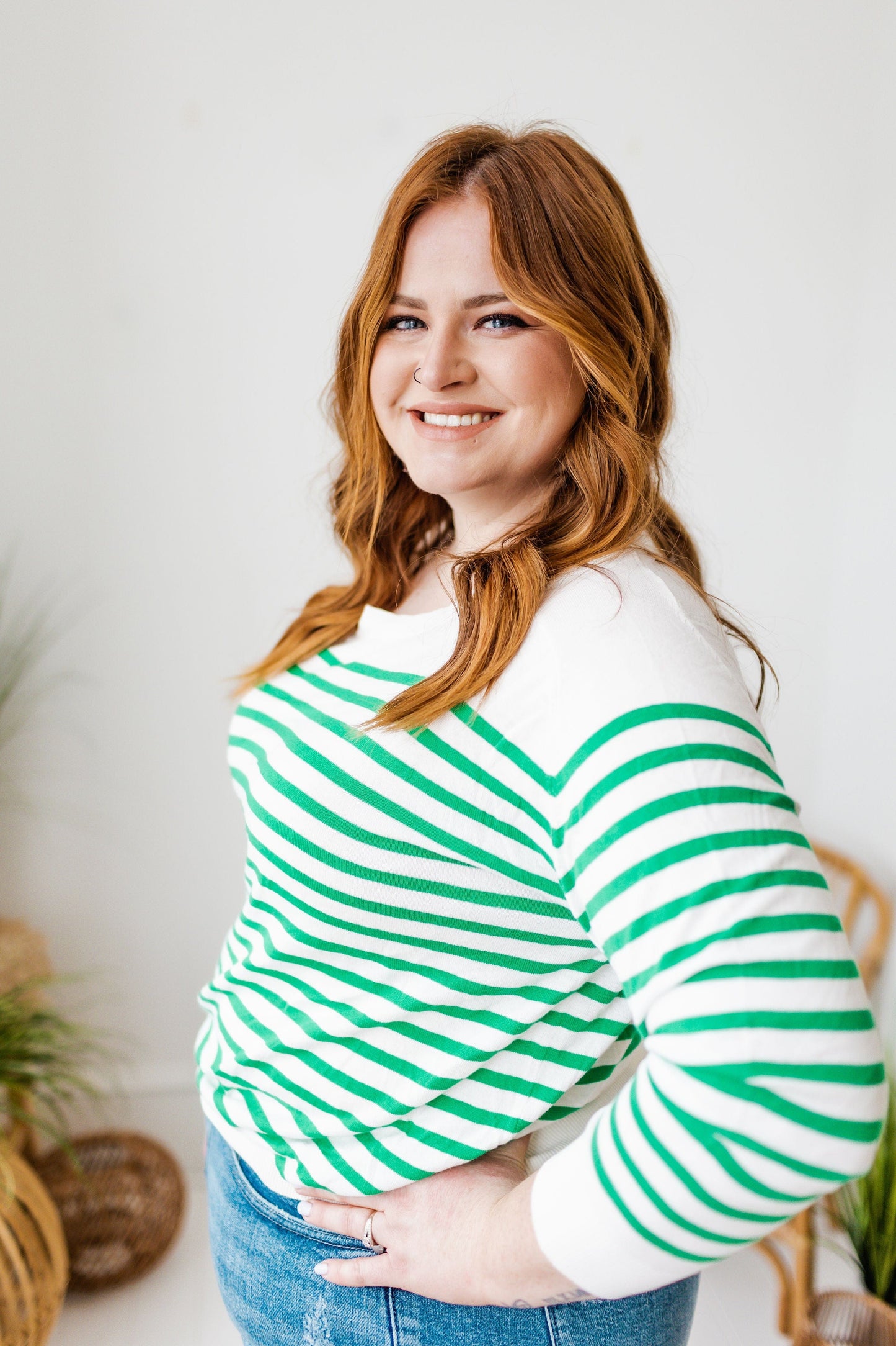 Smiling woman with long hair wearing a classic fit, green and white striped lightweight sweater and jeans, standing with hands on hips in a room adorned with wicker chairs and plants.