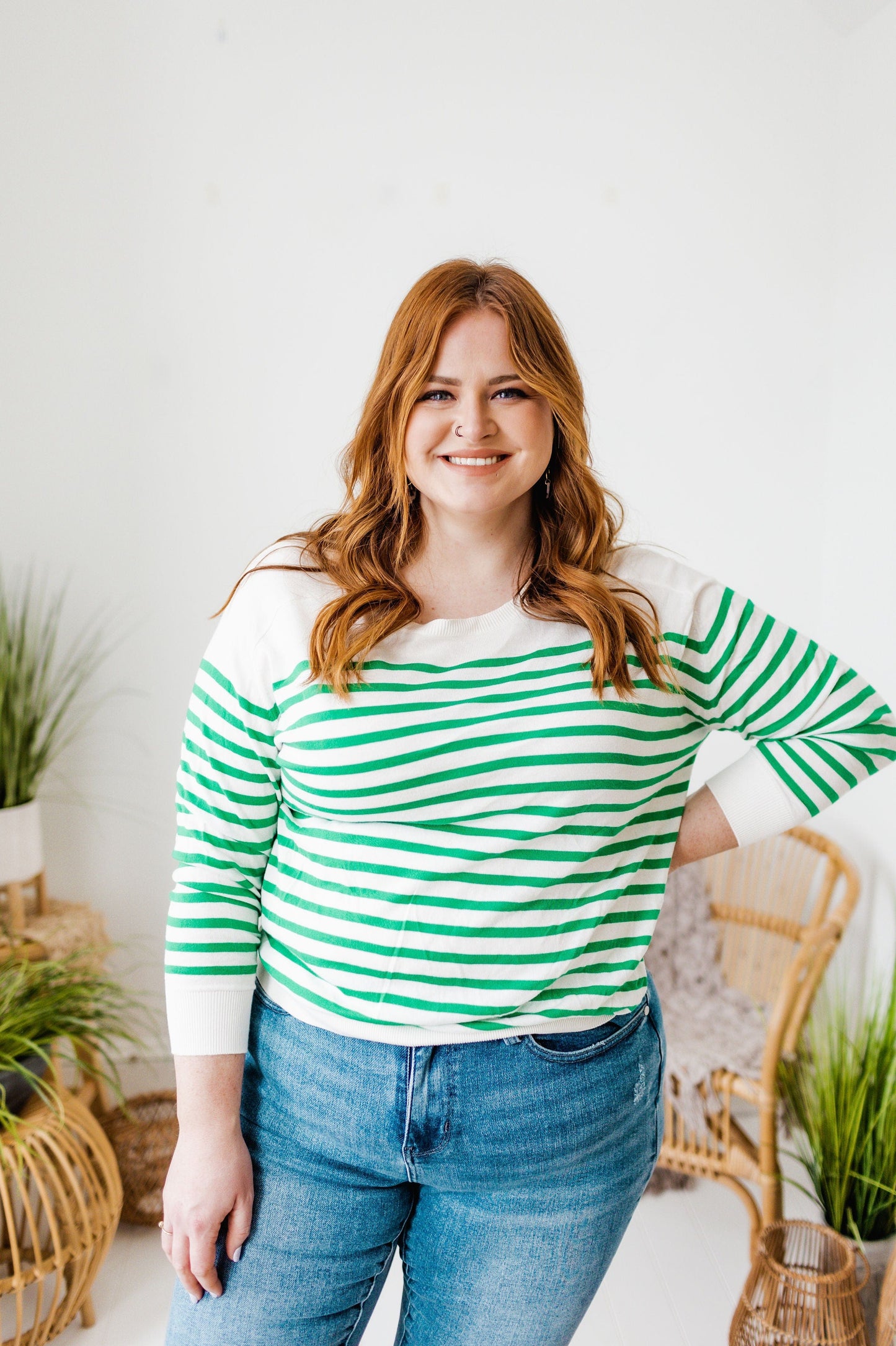 A person with long hair smiles, wearing a classic fit lightweight sweater featuring green stripes and jeans. Wicker chairs and potted plants adorn the background.