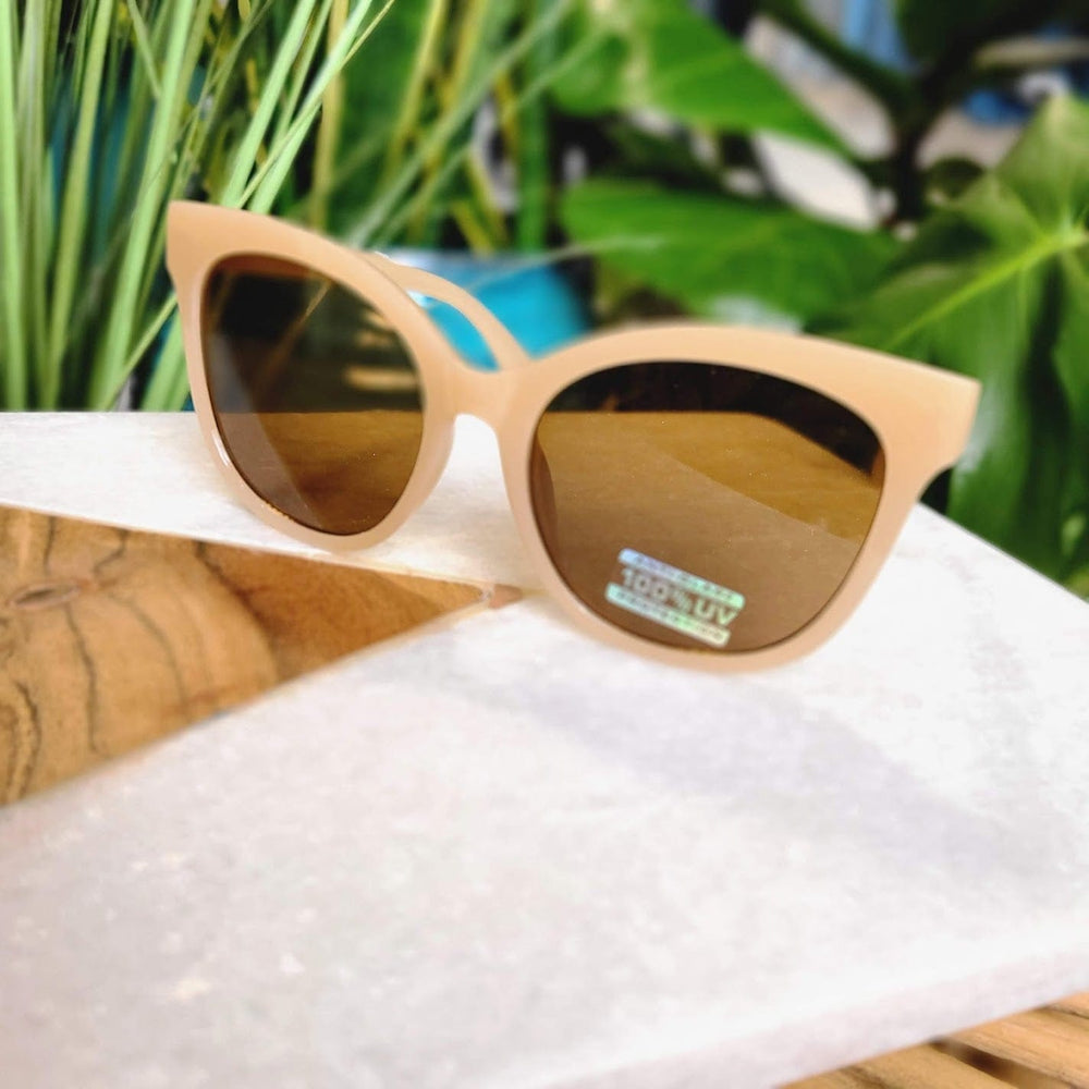 Beige sunglasses with dark lenses rest stylishly on a white and wooden surface, surrounded by lush green plants. These chic sunglasses offer complete UVA/UVB protection and include free shipping for a limited time.
