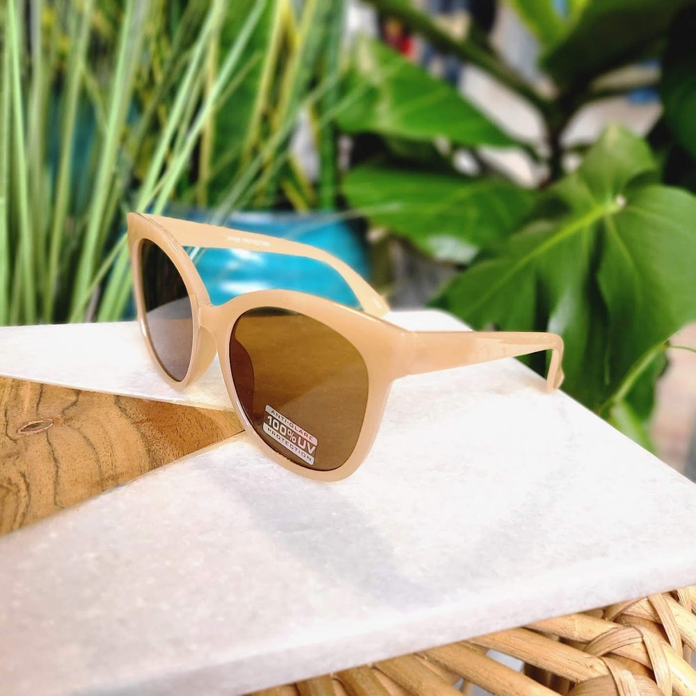 Beige sunglasses with UVA/UVB protection and dark lenses sit on a marble and wood surface, surrounded by lush green leaves.