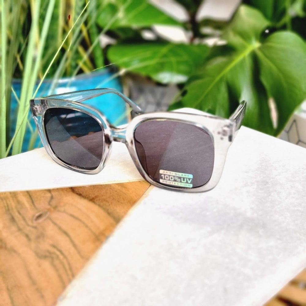 Clear-framed sunglasses in a translucent smoke color, featured with dark lenses and a 100% UVA/UVB protection sticker, elegantly rest on a table surrounded by lush greenery.