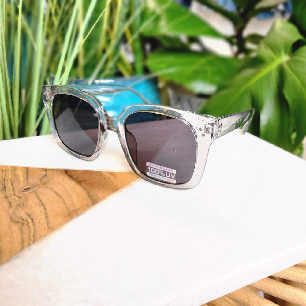 A pair of square-framed sunglasses in a translucent smoke color with a UVA/UVB protection sticker rests on a white and wooden surface, surrounded by vibrant green plants.