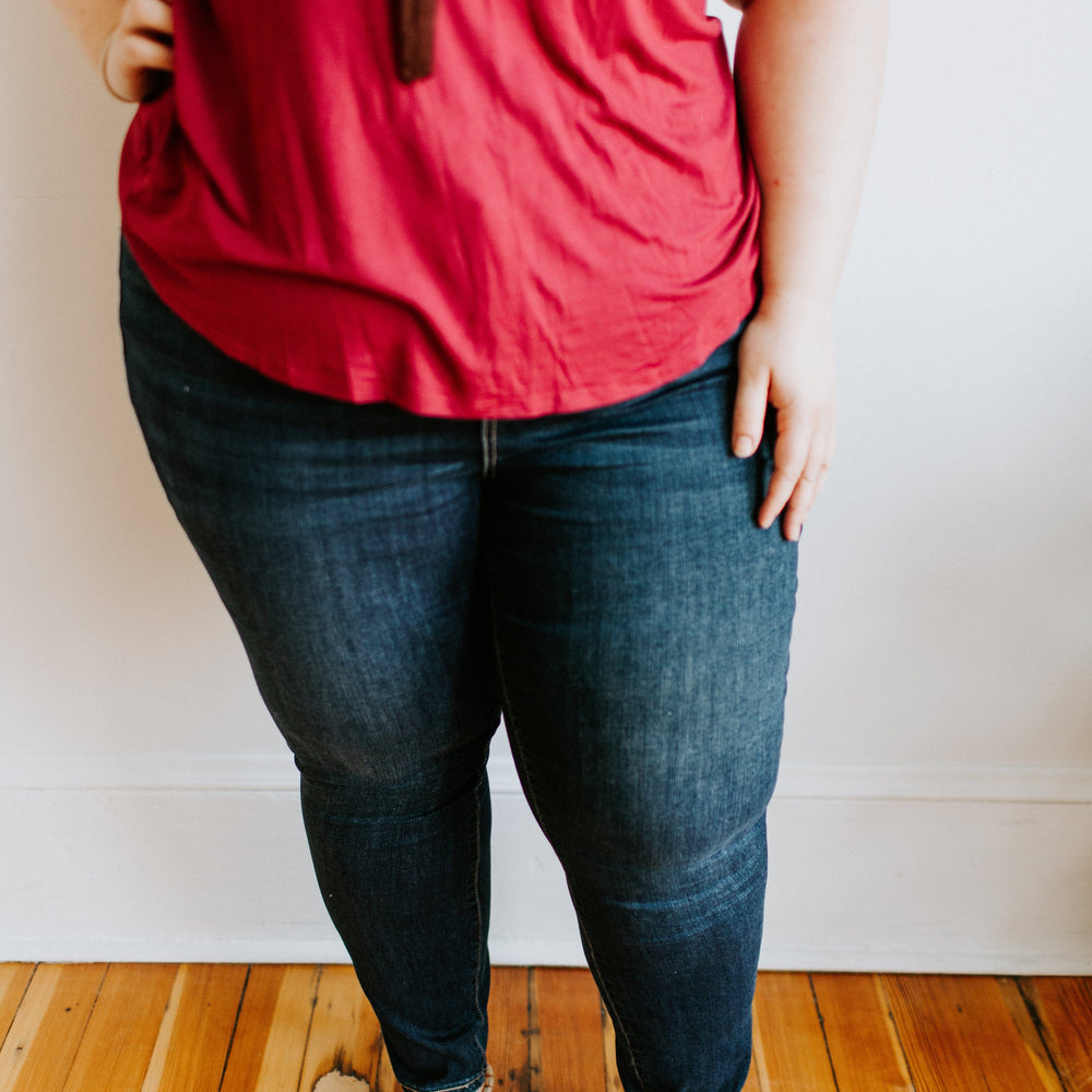 A person dressed in a red shirt and white sandals stands with one hand on their hip, showcasing the Love Marlow MID-RISE SKINNY JEAN WITH ZIPPER DETAIL IN INDIGO on a wooden floor.
