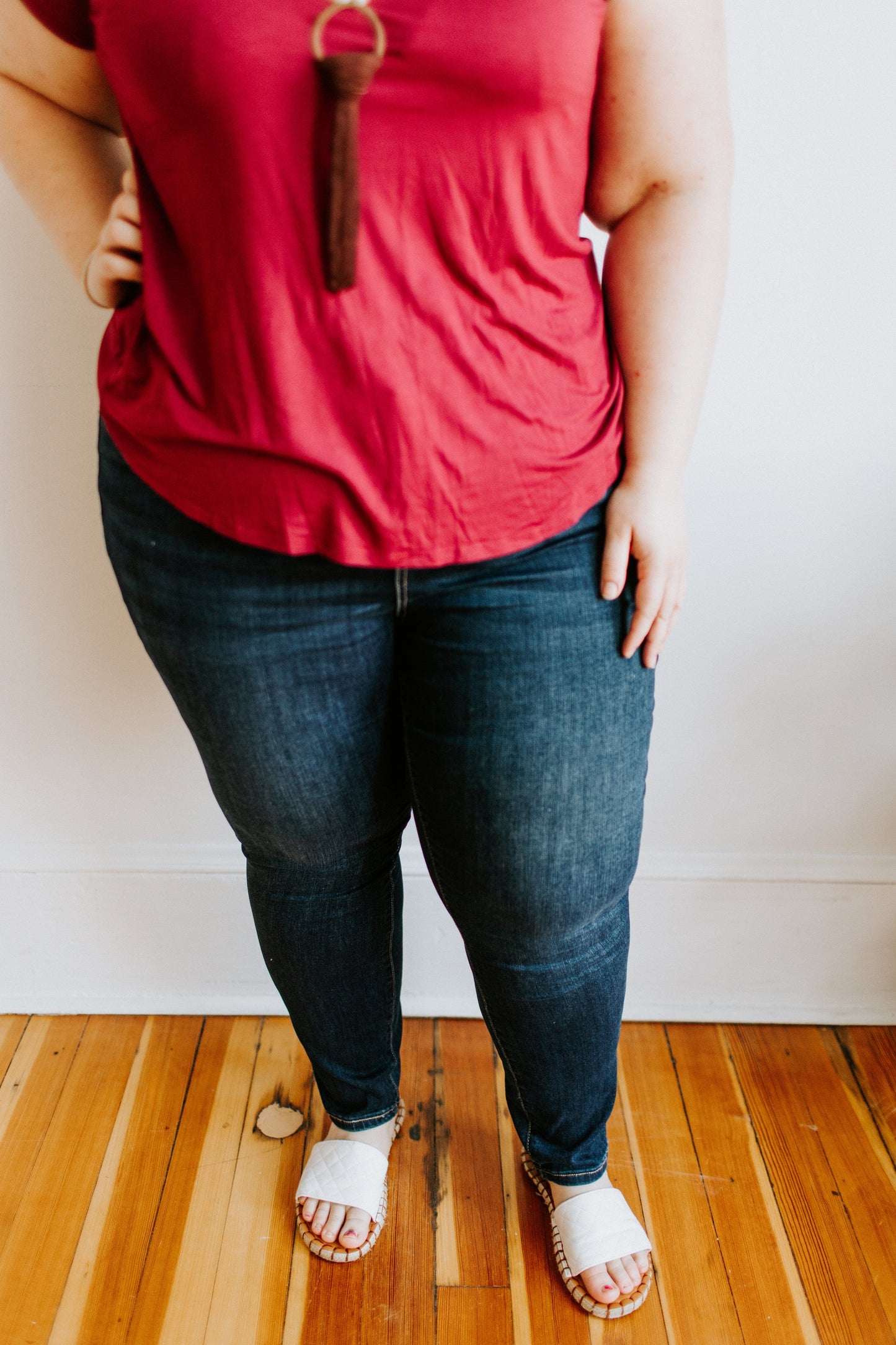 A person dressed in a red shirt and white sandals stands with one hand on their hip, showcasing the Love Marlow MID-RISE SKINNY JEAN WITH ZIPPER DETAIL IN INDIGO on a wooden floor.