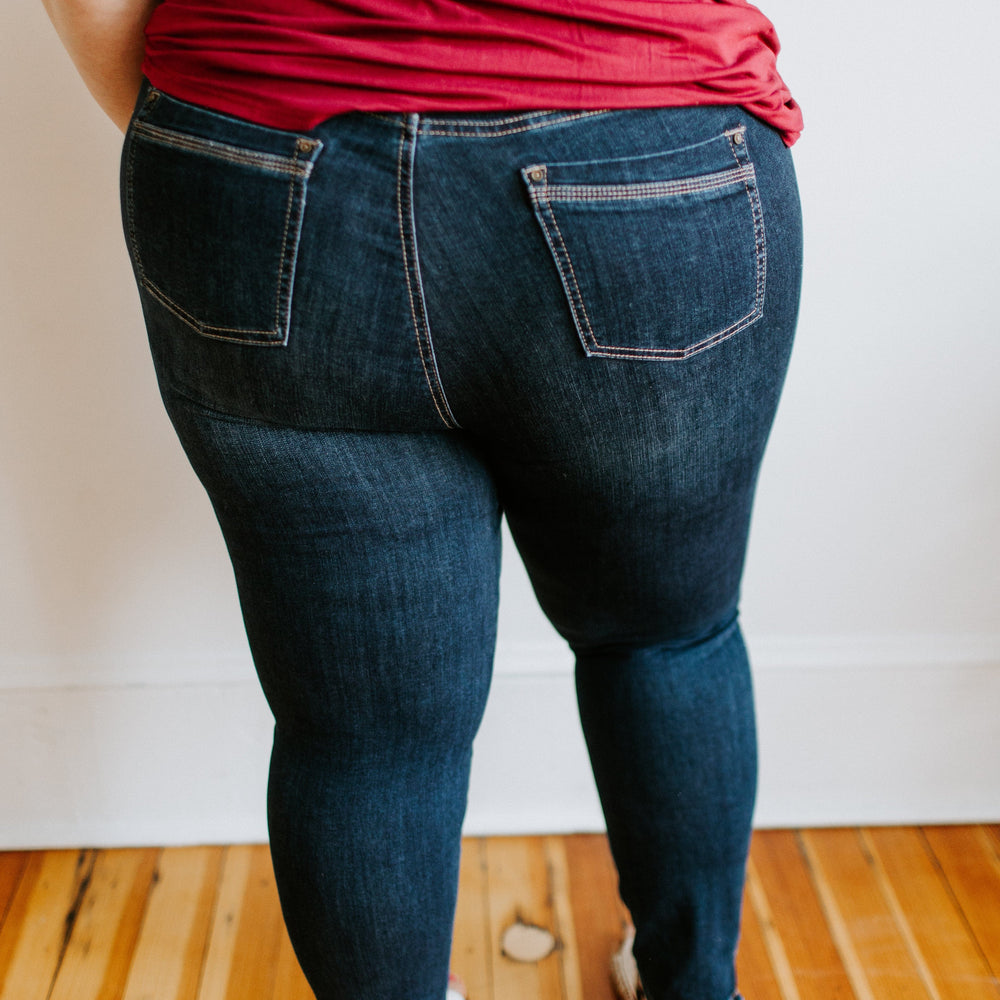 A person in a red shirt and Love Marlow's MID-RISE SKINNY JEAN WITH ZIPPER DETAIL IN INDIGO stands on a wooden floor, facing away from the camera.