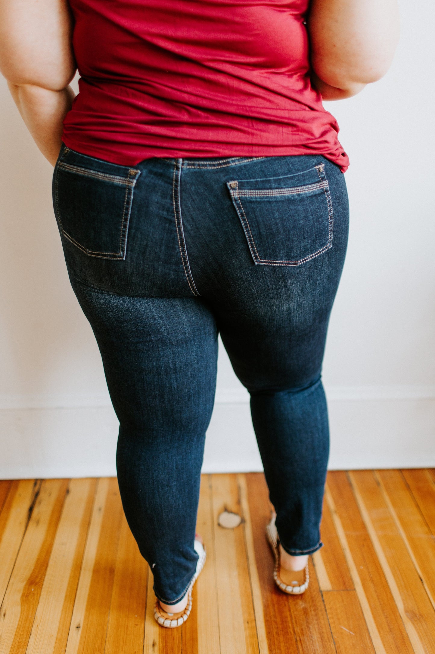 A person in a red shirt and Love Marlow's MID-RISE SKINNY JEAN WITH ZIPPER DETAIL IN INDIGO stands on a wooden floor, facing away from the camera.