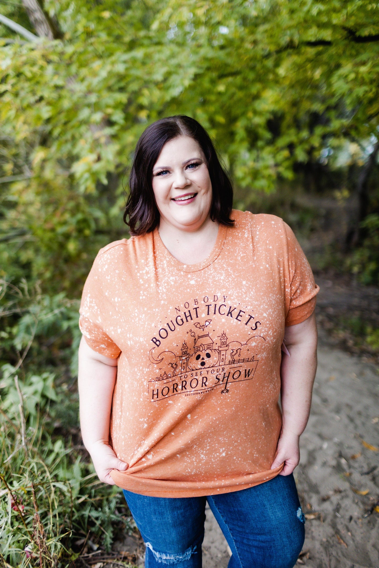 A person wearing the Love Marlow "NOBODY BOUGHT TICKETS TO SEE YOUR HORROR SHOW" graphic tee stands smiling on a wooded path, the vibrant colors of their hand-bleached shirt adding a touch of fun amidst the natural scenery.
