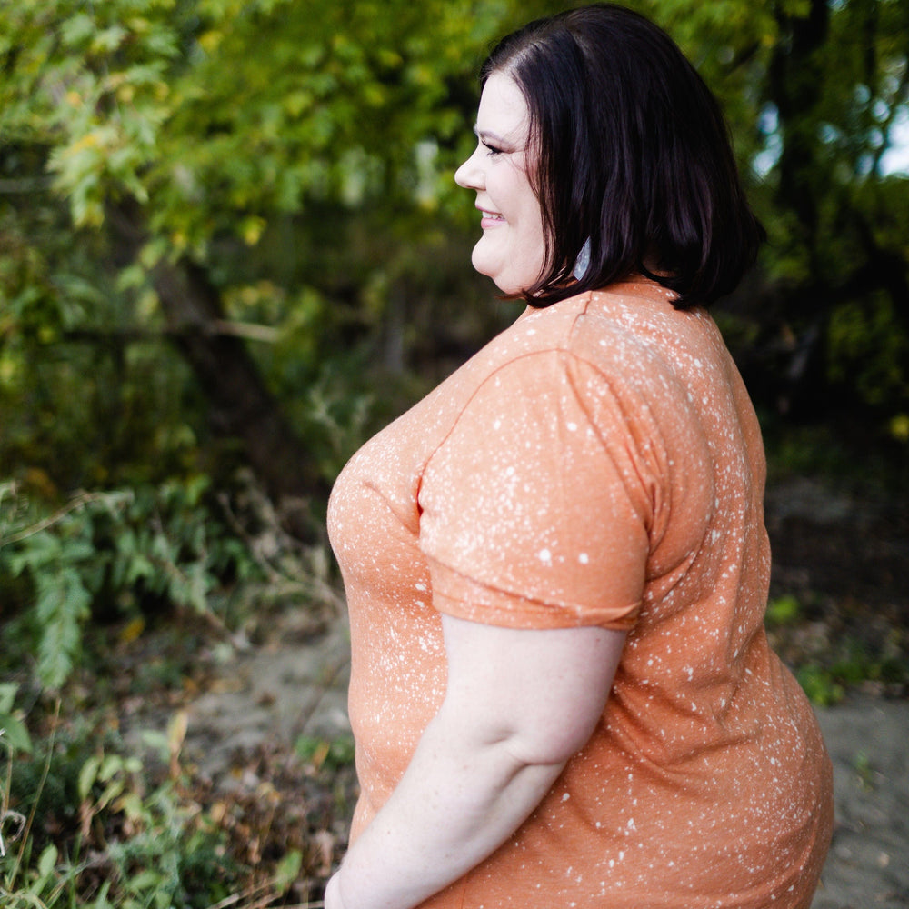 A person stands in profile outdoors, wearing the "NOBODY BOUGHT TICKETS TO SEE YOUR HORROR SHOW" graphic tee by Love Marlow. The shirt's unique orange speckled design, achieved through hand bleaching, is paired with jeans. The vibrant greenery in the background complements this distinct style.