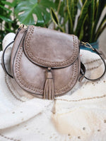 A beige crossbody satchel with decorative stitching opens to reveal a patterned fabric lining, placed on a textured white cloth. The bag features a tassel detail and plants are visible in the background.