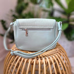 A light blue crossbody satchel rests on a wicker chair, showcasing its plaid interior and zipper. The bag, crafted from vegan-faux leather, is elegantly complemented by woven straw detail.