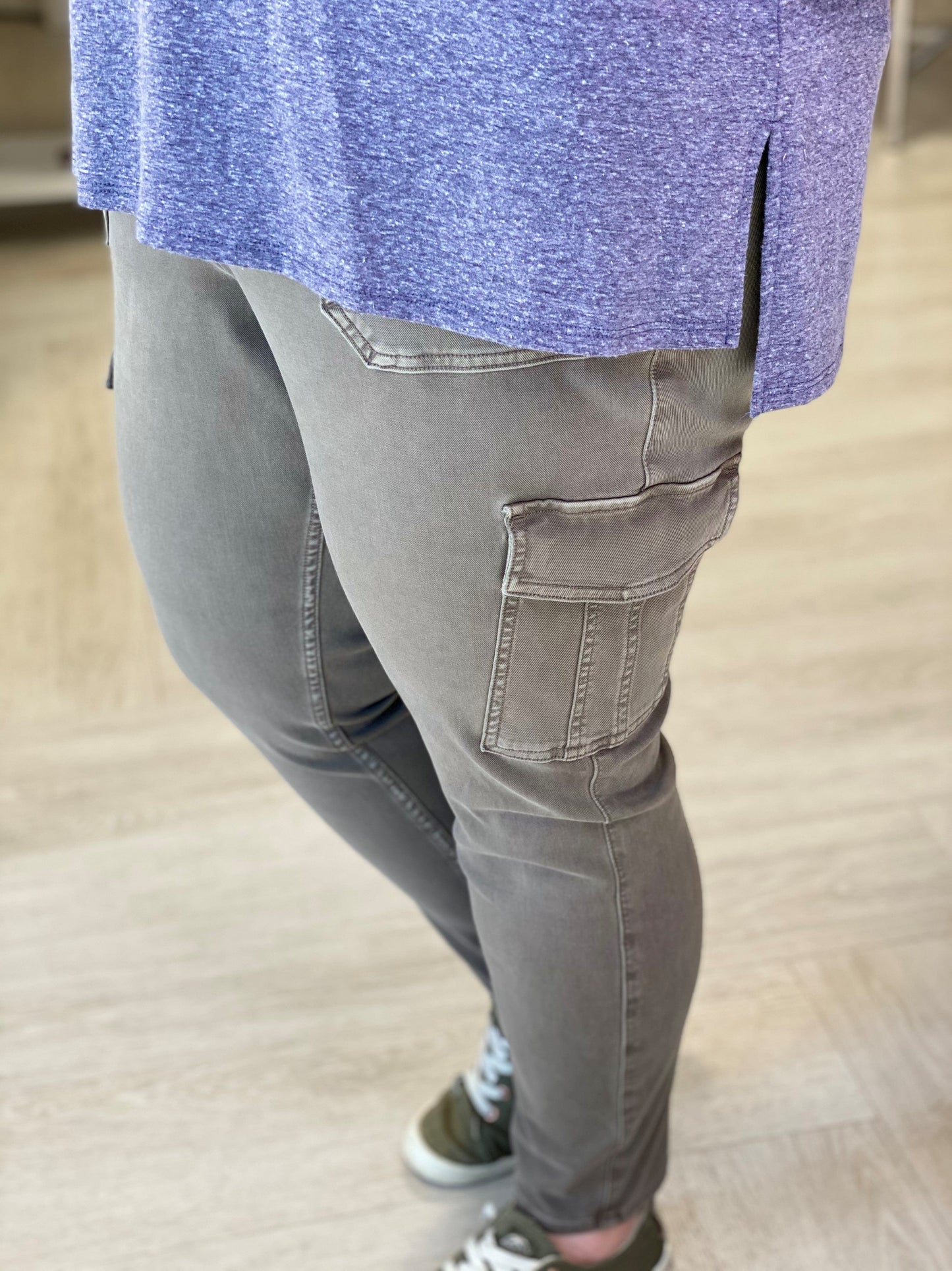 A person wearing Spanx® Stretch Twill Ankle Cargo Pants in Desert Dune with hidden Gut Check™ shaping and green sneakers stands on a light wood floor.