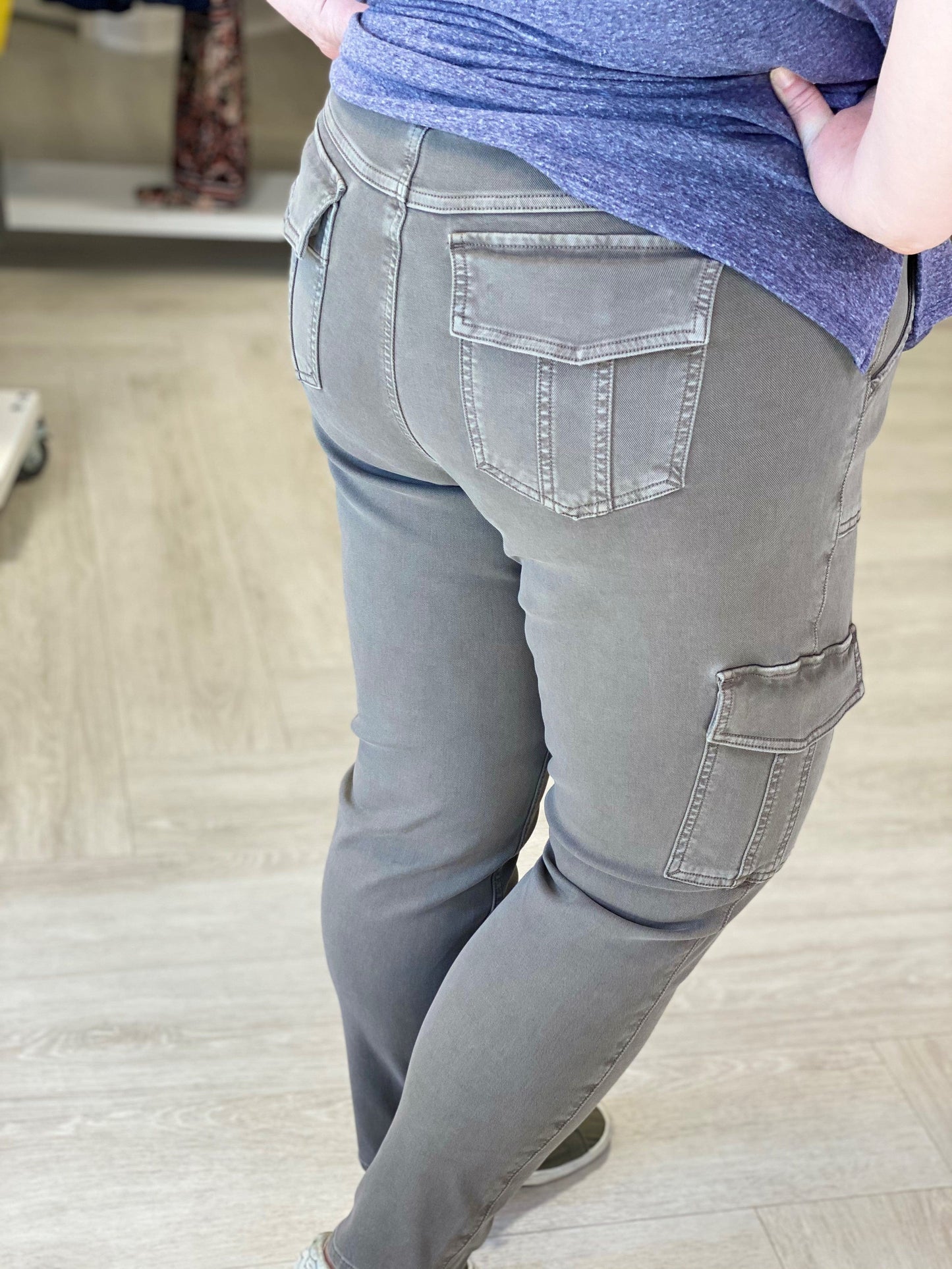 The individual is dressed in the comfortable Spanx® Stretch Twill Ankle Cargo Pant in Desert Dune and a blue shirt, standing indoors on a light wood floor. These gray cargo pants feature hidden Gut Check™ shaping for both comfort and style.