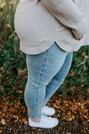 A person in a beige sweater and Spanx© ANKLE SKINNY JEANS by Love Marlow, made from comfortable stretch denim in light blue, stands on a leaf-covered path wearing white sneakers.