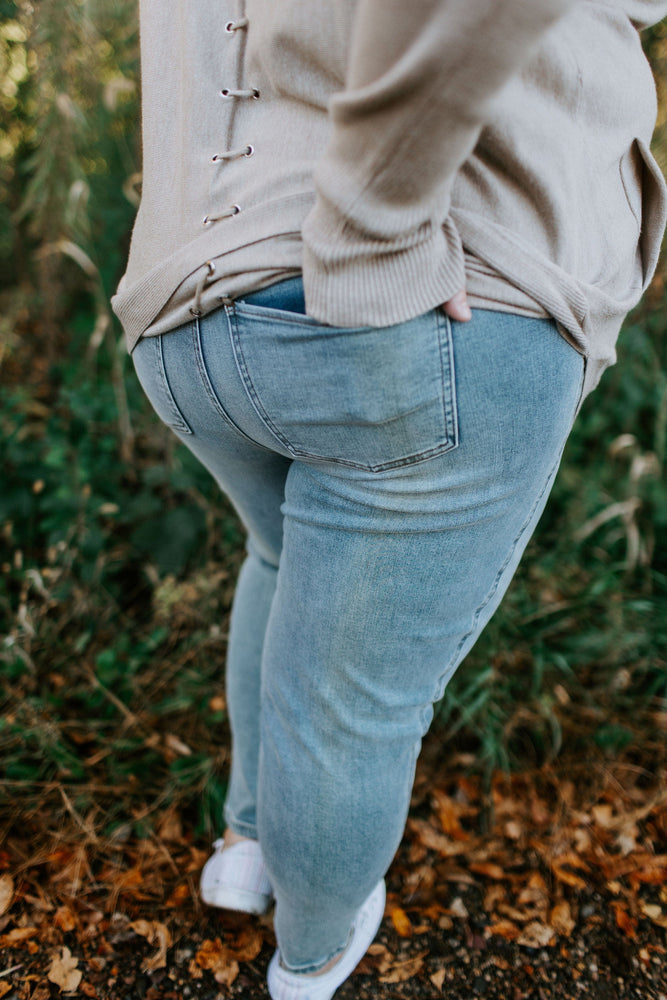 Someone wearing Love Marlow's Spanx© ANKLE SKINNY JEANS and a beige sweater stands outdoors on a leafy path, relishing the comfort of high-rise coverage.