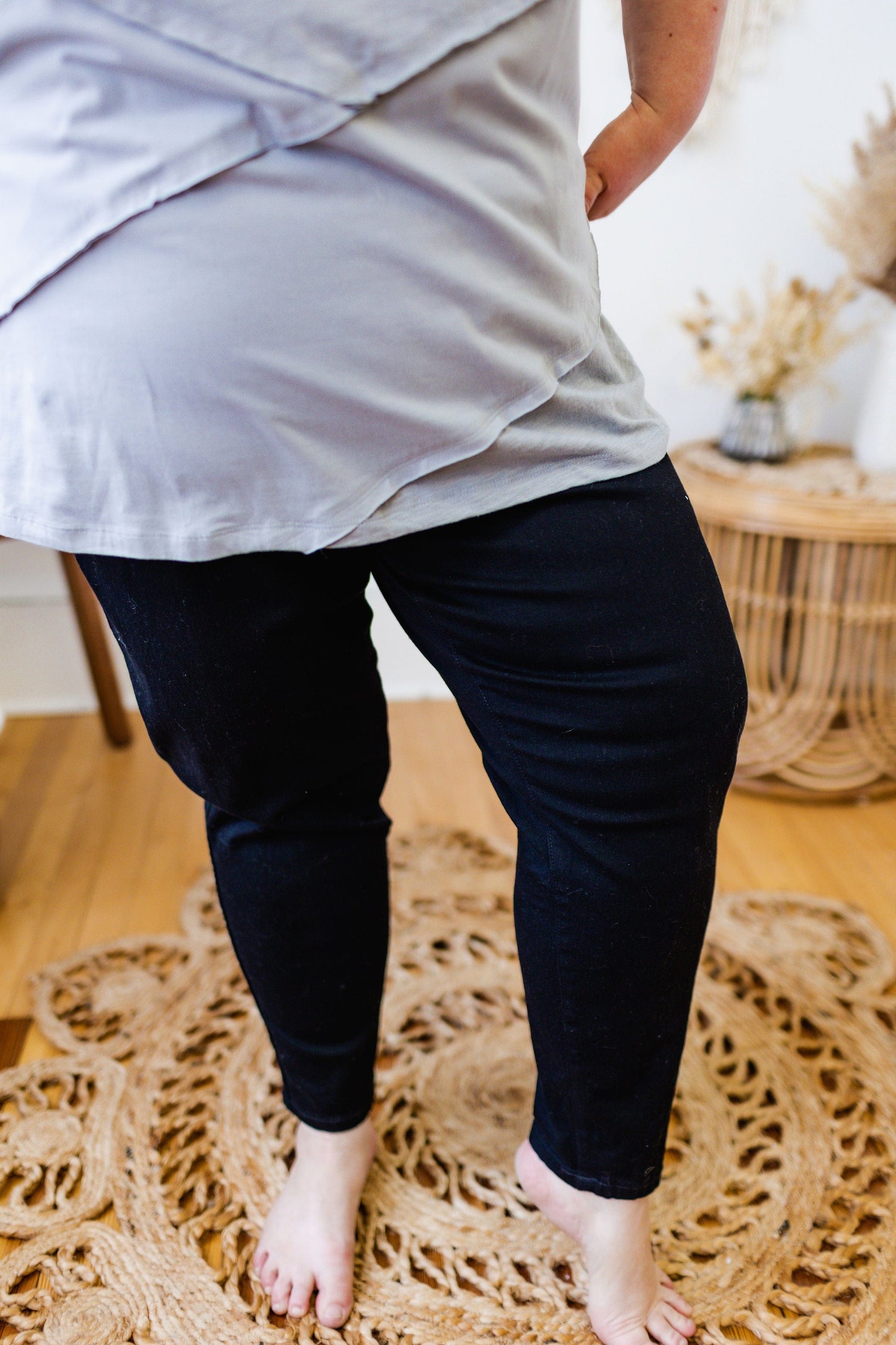 A person in a light shirt and Spanx© Ankle Skinny Jeans in black from Love Marlow stands on a woven rug in a room with a wooden floor and wicker furniture.