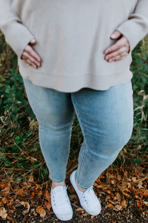 A person in a beige sweater and Love Marlow's comfortable Spanx© ANKLE SKINNY JEANS in light blue stands outdoors on a leafy ground, with hands resting on their hips.