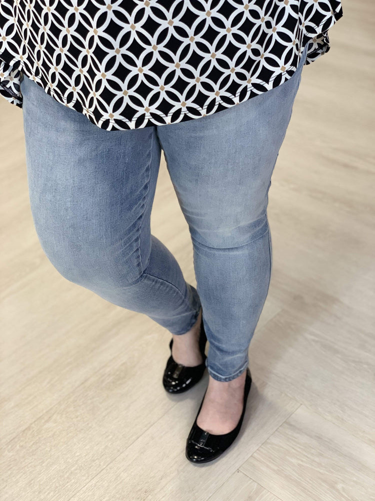 A person stands on a light wood floor, wearing Spanx© Ankle Skinny Jeans from Love Marlow made of comfortable stretch denim, paired with a black and white patterned top and black shoes.