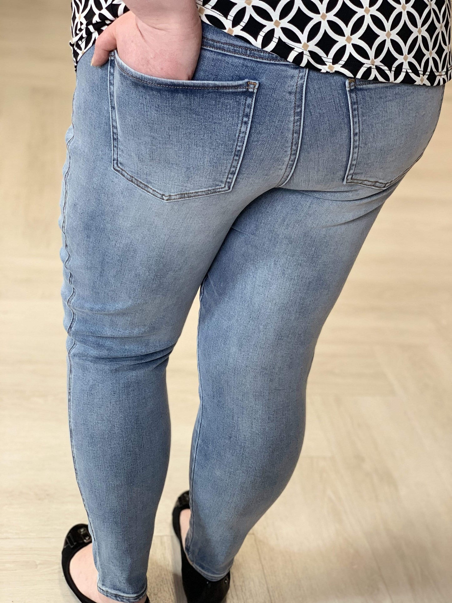 A person wearing Love Marlow's Spanx© Ankle Skinny Jeans and a patterned black and white top, standing on a light wood floor.