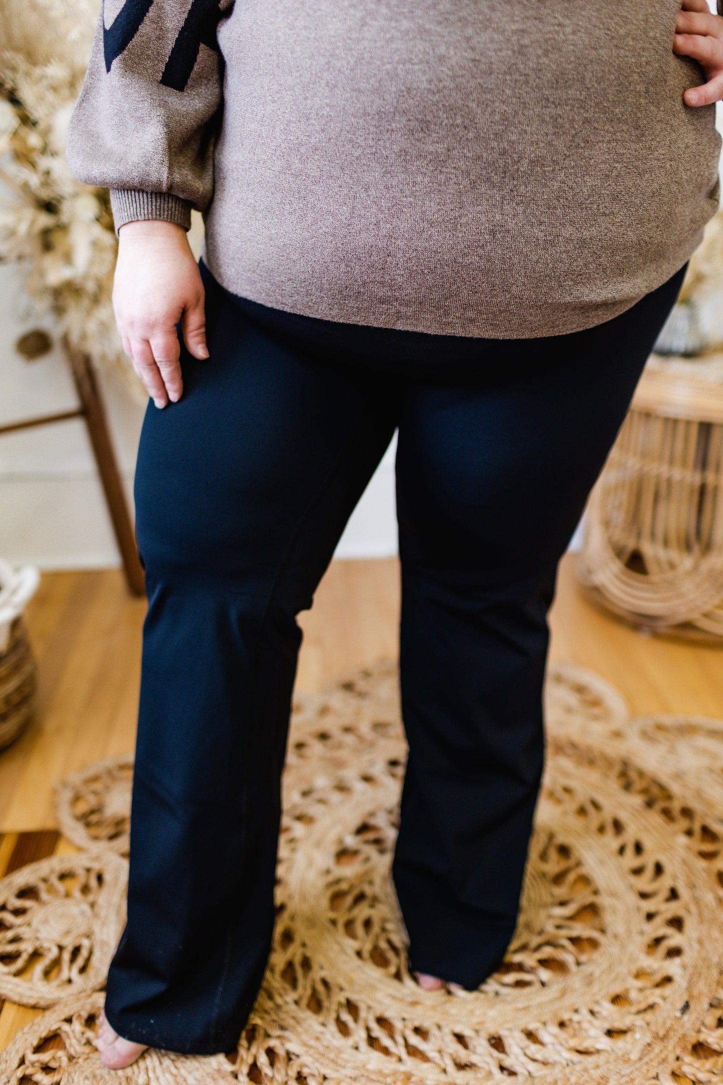 A person stands on a woven rug, wearing a brown sweater and the Love Marlow Spanx© BOOTY BOOST YOGA PANT, crafted from sculpting performance fabric and featuring a contoured waistband.