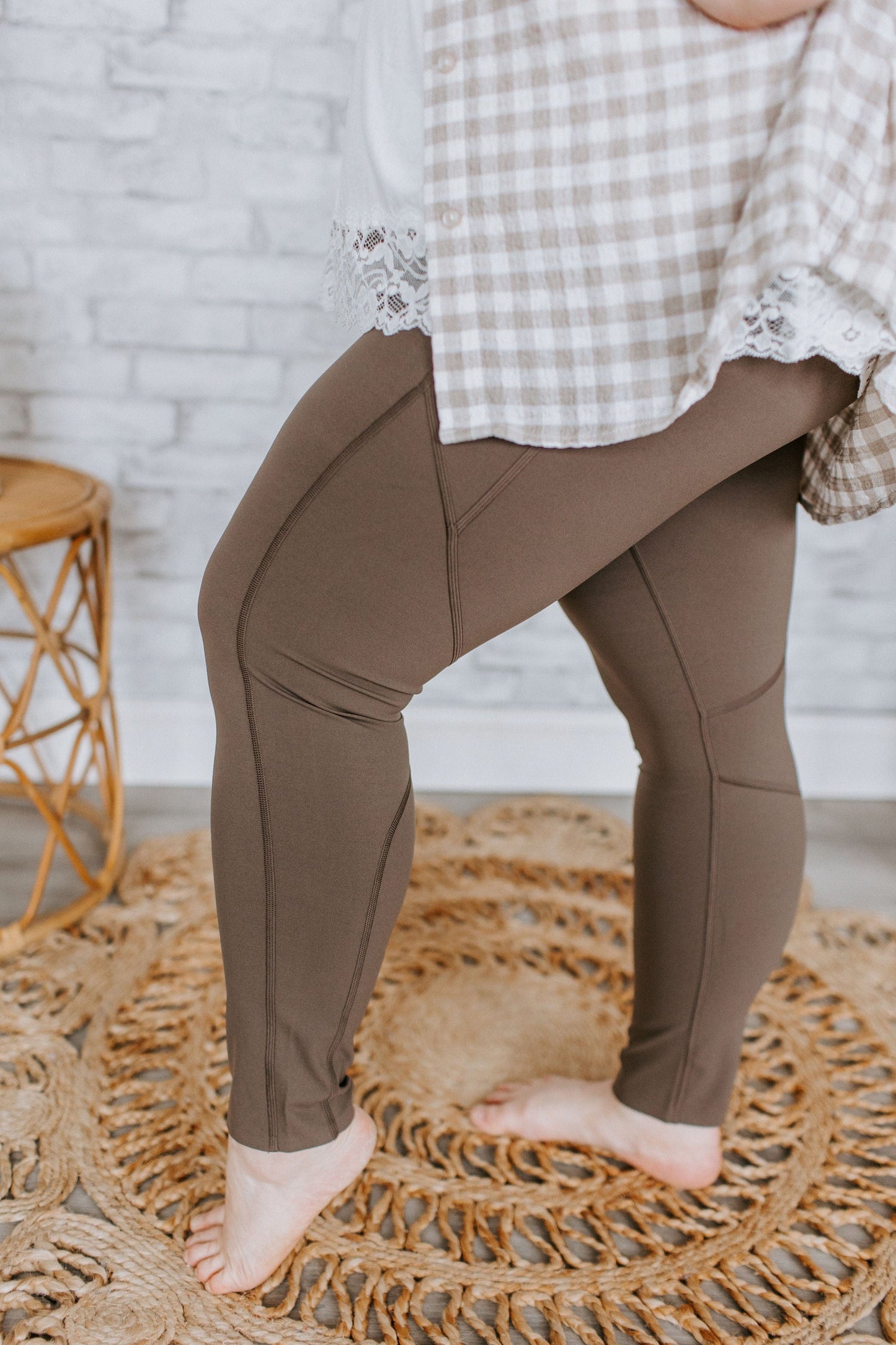 A person stands barefoot on a woven rug, wearing Spanx© EVERY.WEAR KNOCKOUT LEGGINGS IN BARK and a plaid shirt with lace trim. Next to them is a wicker stool by a light-colored brick wall.