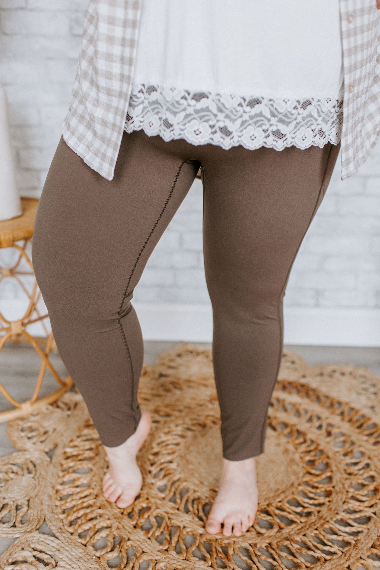 A person wearing Spanx© EVERY.WEAR KNOCKOUT LEGGINGS IN BARK, a white top with lace trim, and a checkered shirt, stands barefoot on a woven rug.