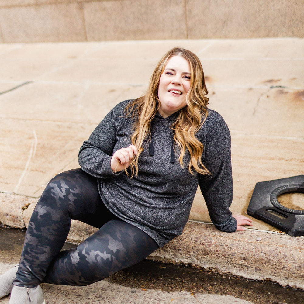 A person with long hair sits on a sidewalk, wearing a gray hoodie, Spanx© Faux Leather Camo Leggings with a Power Waistband, and gray shoes, smiling at the camera.