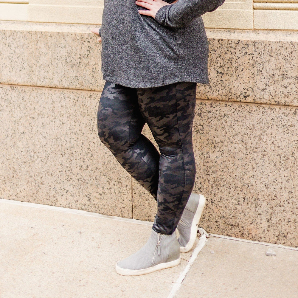A person wearing a gray sweater, Spanx© FAUX LEATHER CAMO LEGGING with a Power Waistband, and gray ankle boots stands confidently with one hand on their hip in front of a beige stone wall.