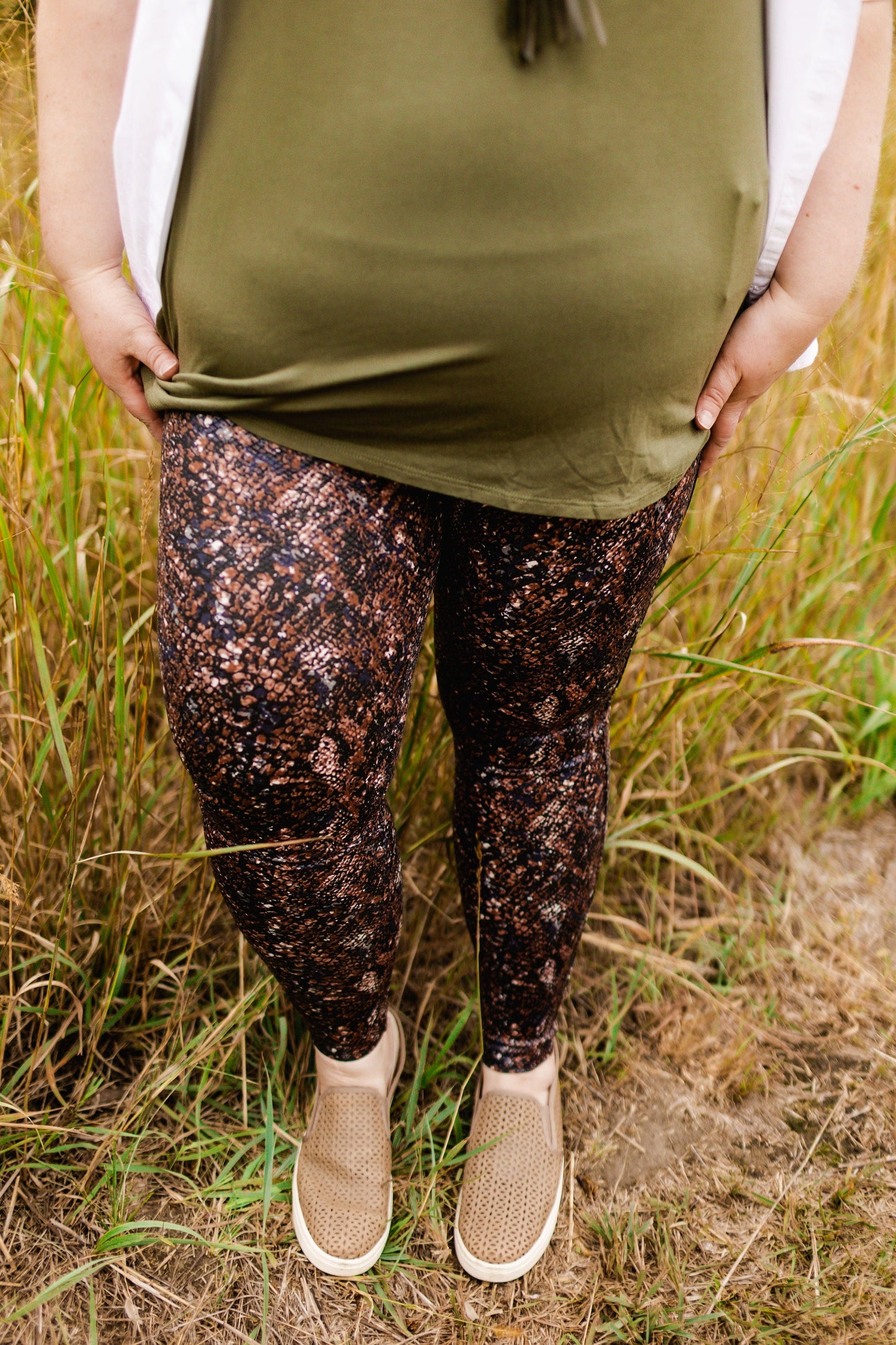 A person is standing in a field, wearing an olive green top paired with Spanx FAUX LEATHER SNAKE SHINE LEGGINGS IN MOCHA SNAKE, featuring a power waistband, along with slip-on shoes. Only the lower half of their body is visible.