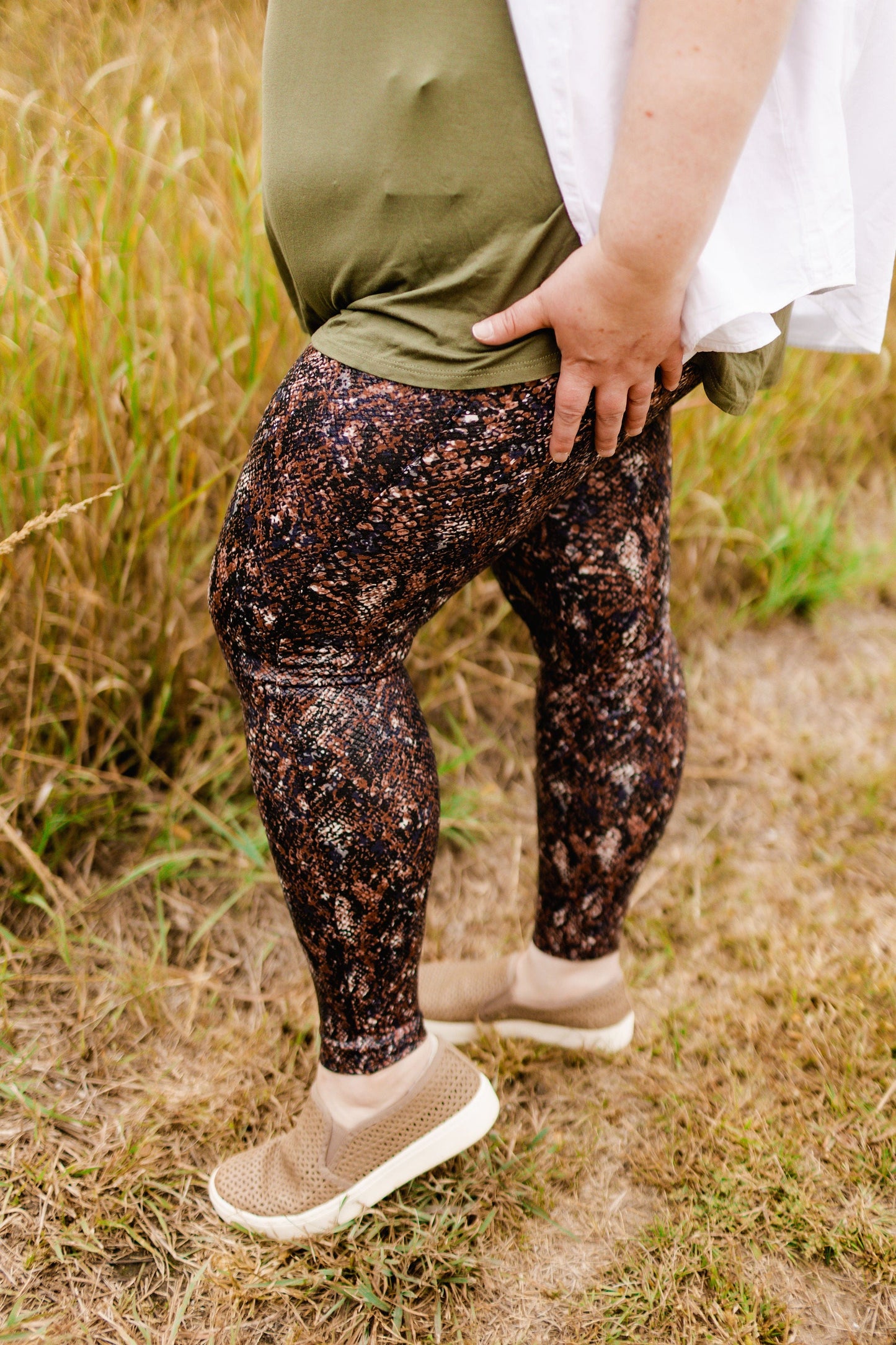 A person wearing Spanx's Faux Leather Snake Shine Leggings in Mocha Snake, paired with a green shirt and beige shoes, stands outdoors on the grass with their hand confidently resting on their hip.