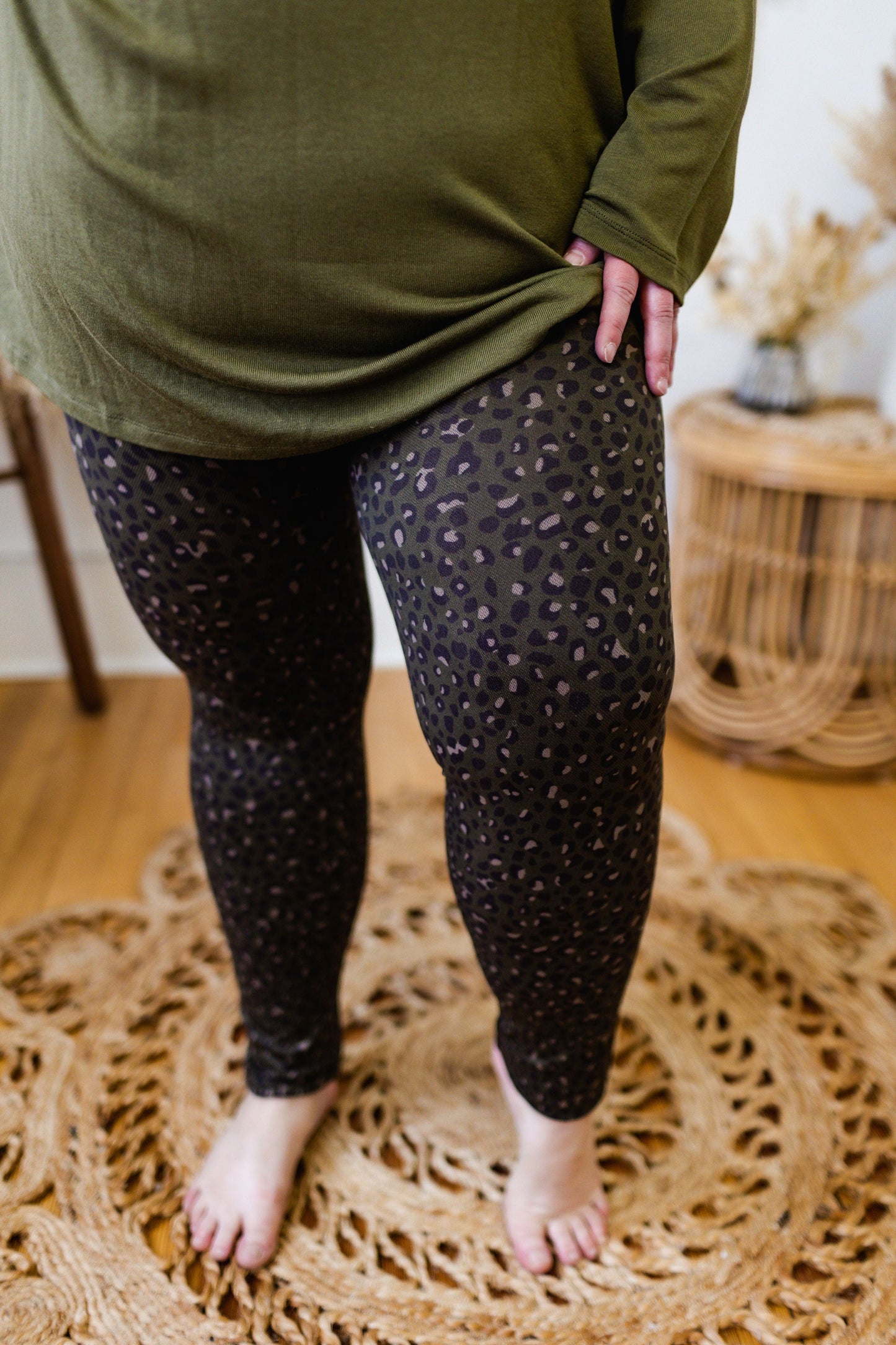 A person wearing Spanx's LOOK AT ME NOW Seamless Leggings in Olive Leopard is standing barefoot on a woven circular rug, while a wicker stool with dried flowers can be seen in the background.