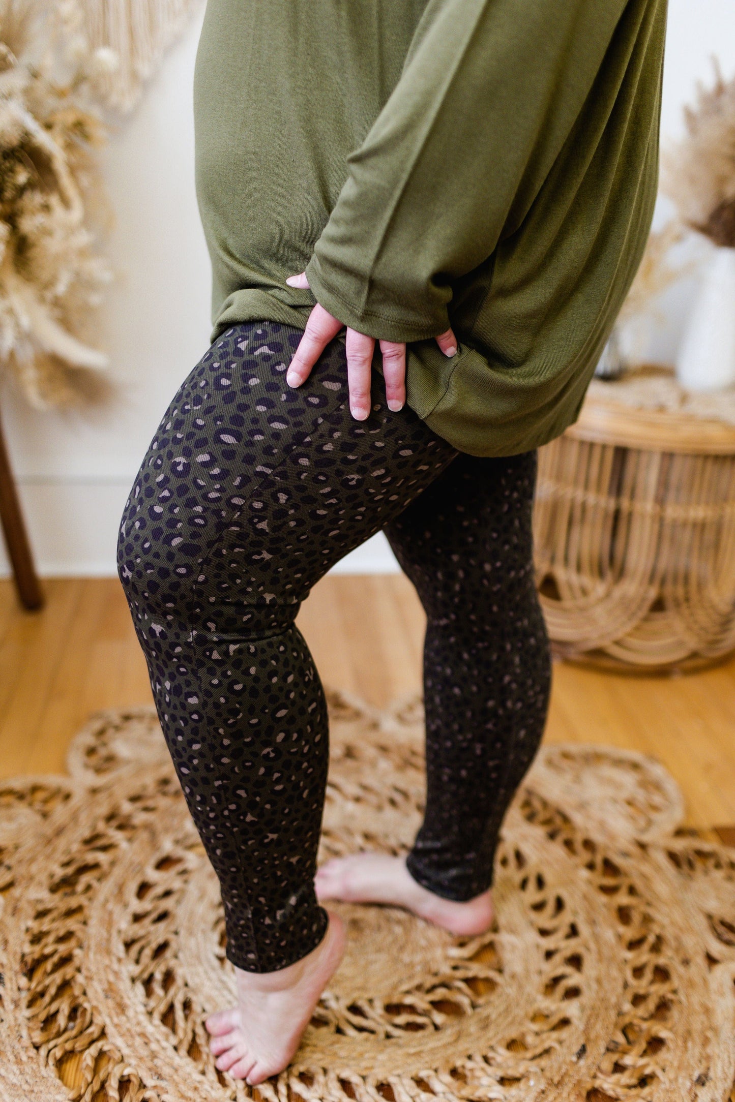 A person wearing a green top and comfortable Spanx© Look At Me Now Seamless Leggings in Olive Leopard stands on a patterned rug, one hand resting on the thigh.