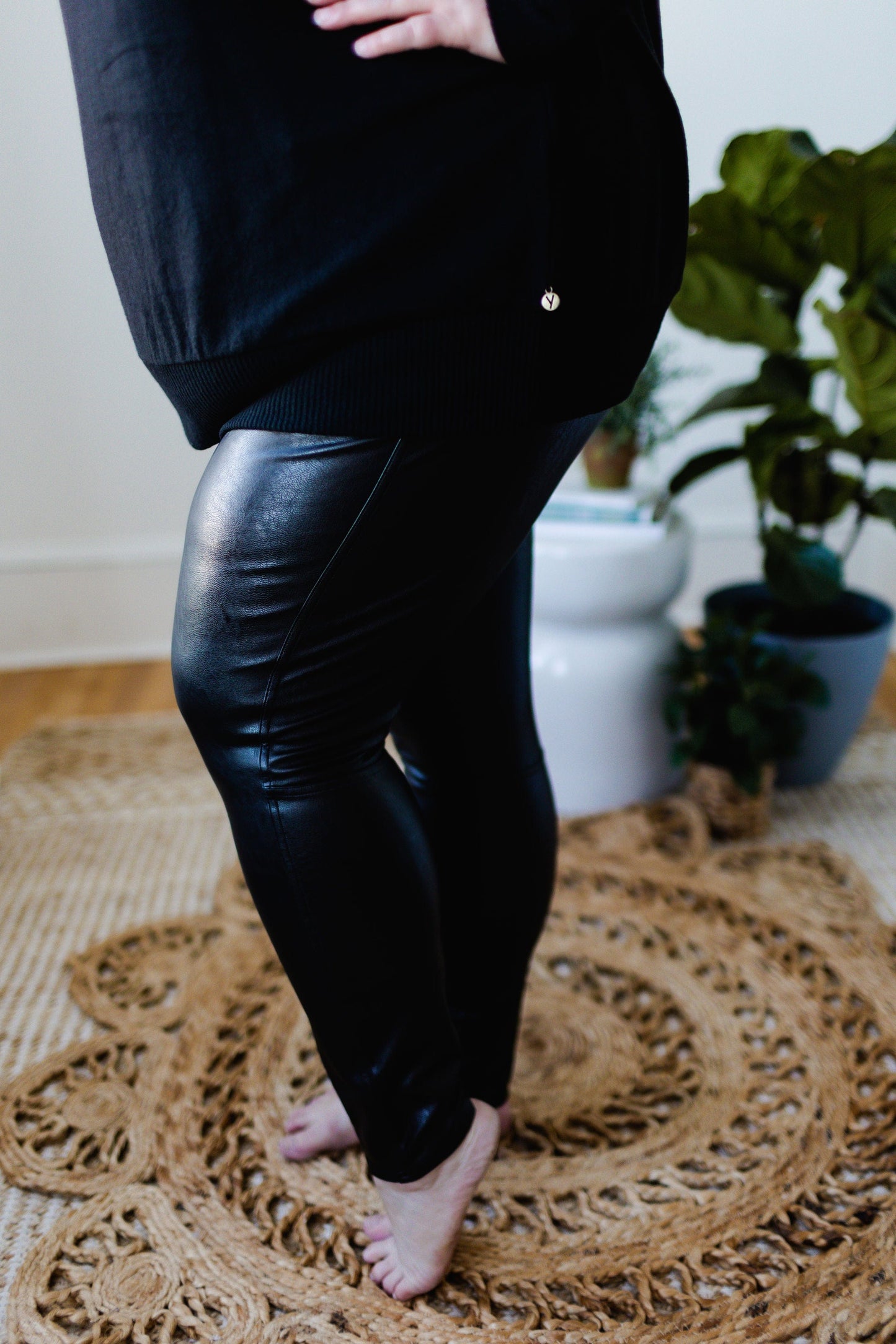 A person stands barefoot on a woven rug, wearing Spanx© Leather-Like Ankle Skinny Pants and a matching top, with a potted plant adding a touch of greenery in the background.